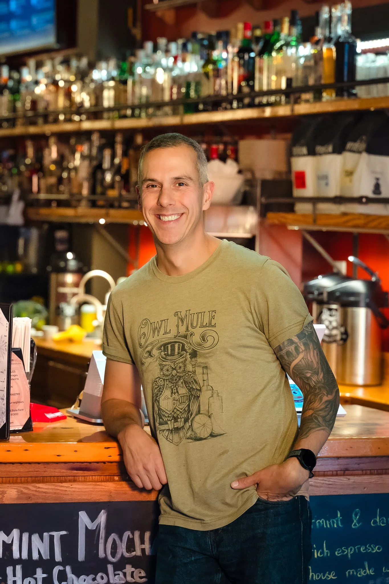 A smiling man with short hair and tattooed arm leaning on a bar counter inside a bar or cafe with bottles of alcohol on shelves in the background.