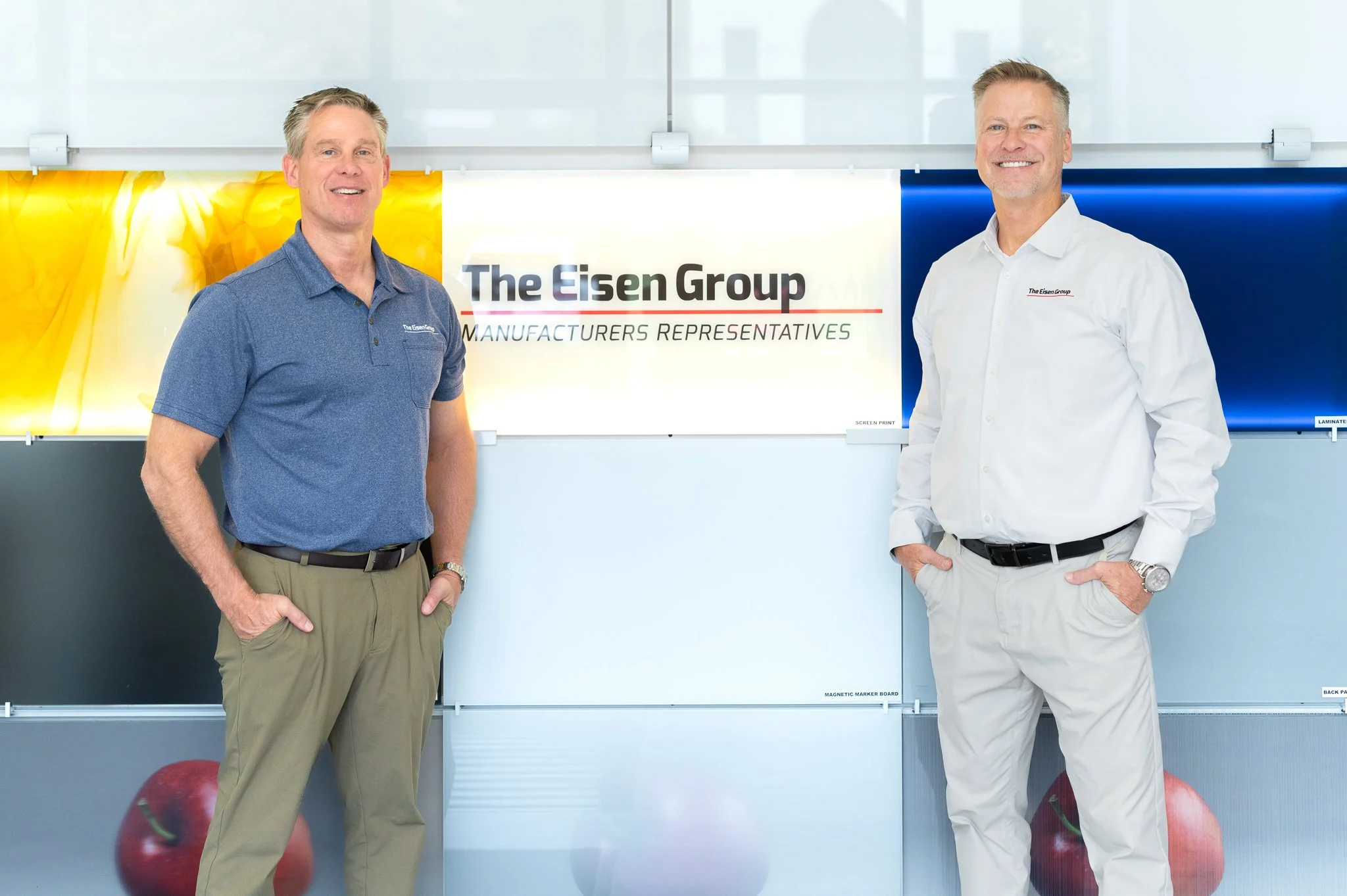 Two men standing indoors in front of a sign that reads "The Eisen Group Manufacturers Representatives". One is wearing a blue polo shirt and khaki pants, the other is wearing a white dress shirt and light-colored pants.