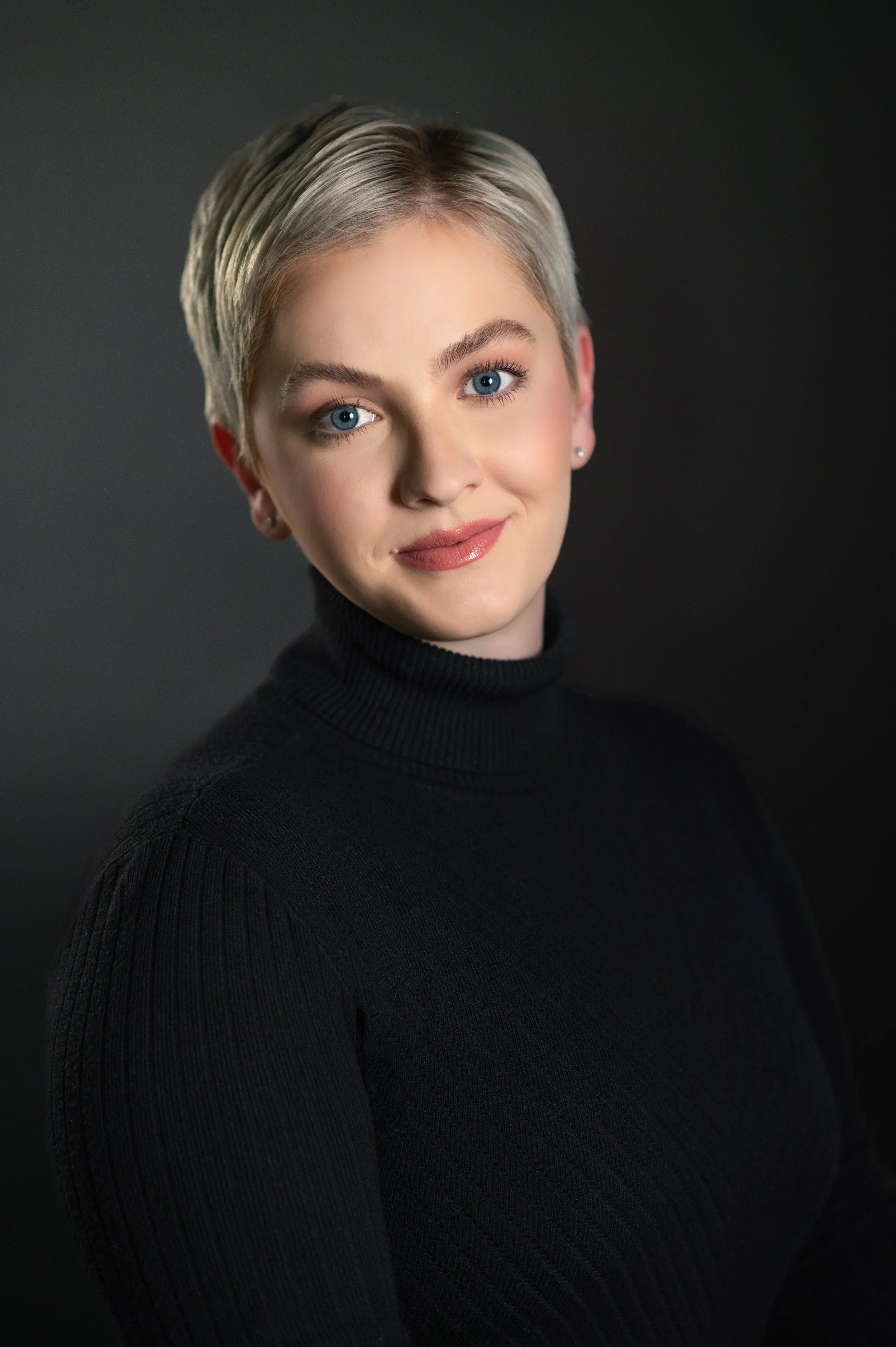 Portrait of a young woman with short blonde hair, blue eyes, wearing a black turtleneck, smiling softly against a dark background.