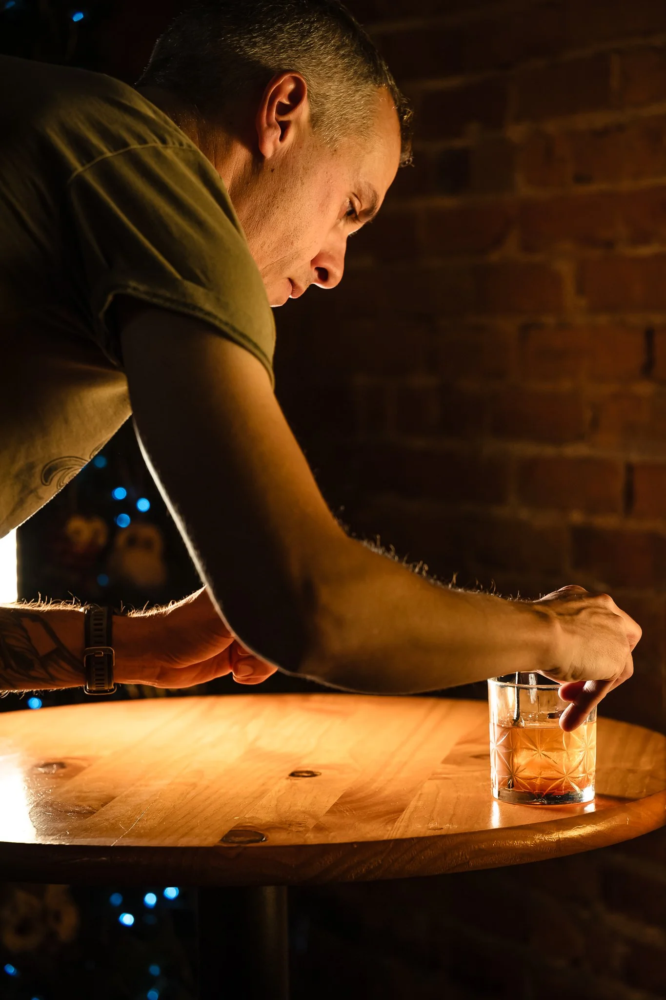 A man is pouring a drink into a glass on a wooden table in a dimly lit setting, with a brick wall background.