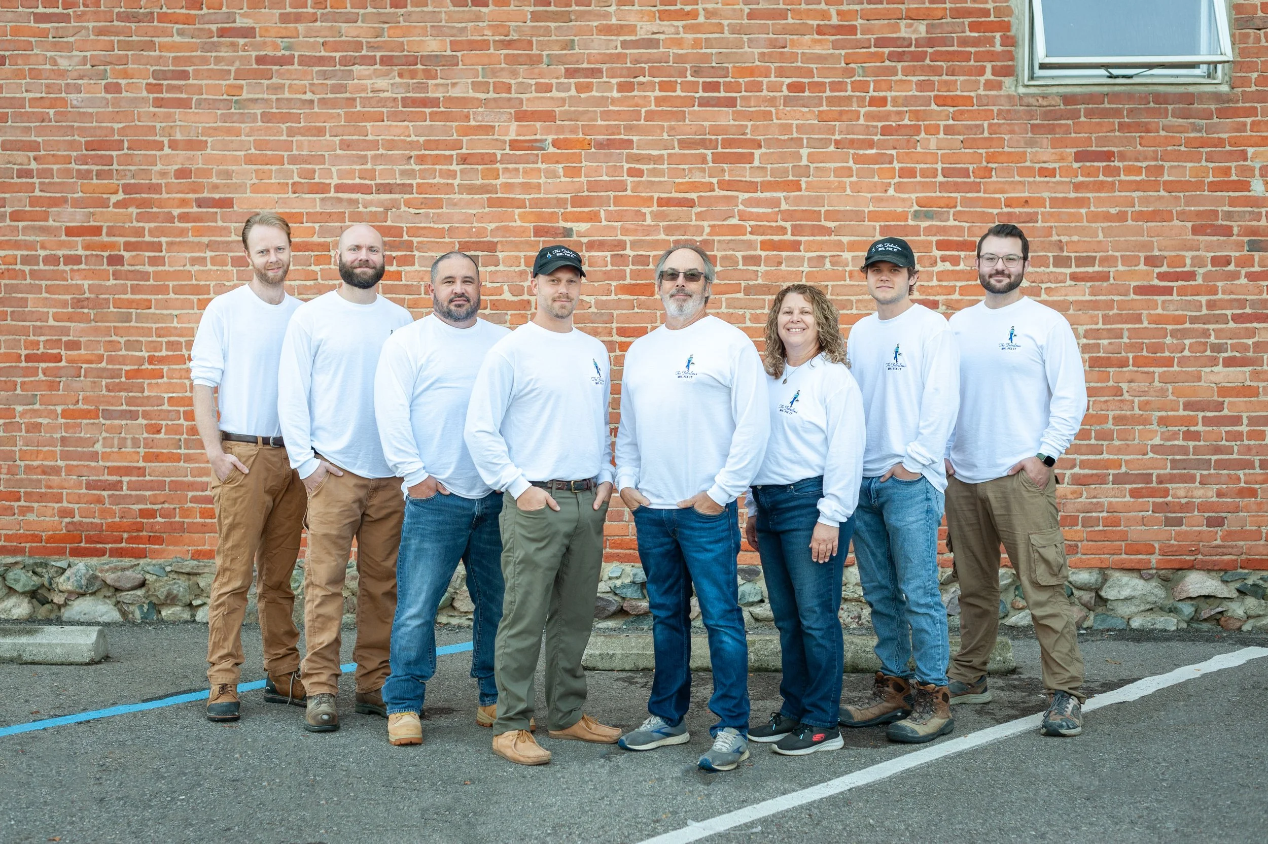 Group of nine people standing in front of a brick wall, all wearing white long-sleeve shirts and casual pants, posing for a photo outside.