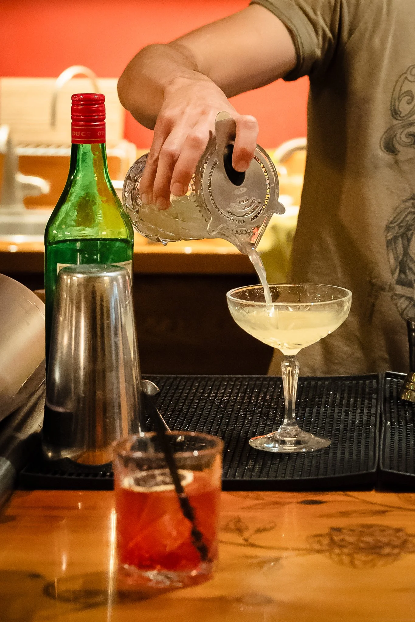 A bartender pouring a cocktail from a shaker into a coupe glass at a bar.