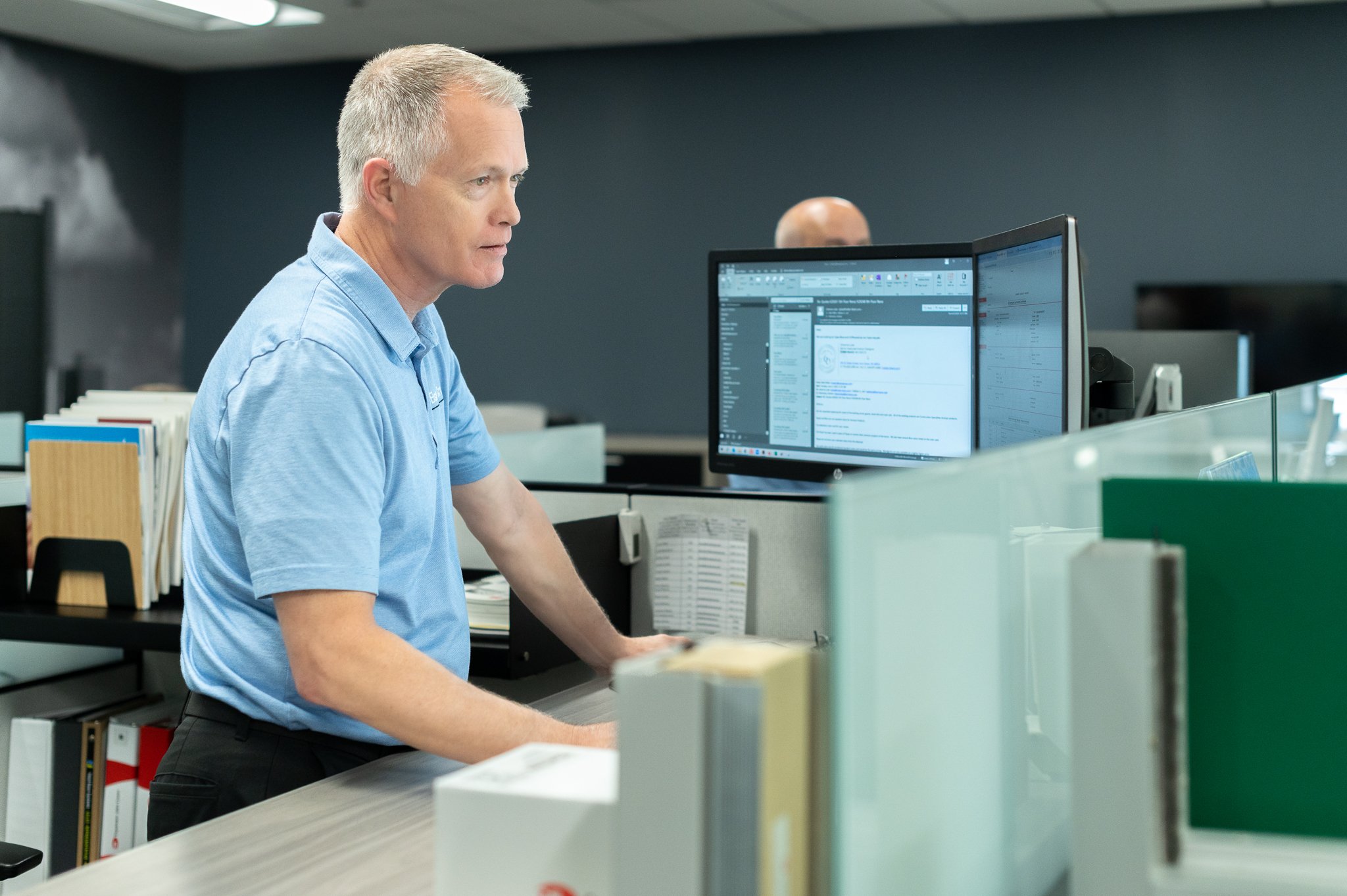 A man in a blue polo shirt working at a desk with dual monitors in an office setting.