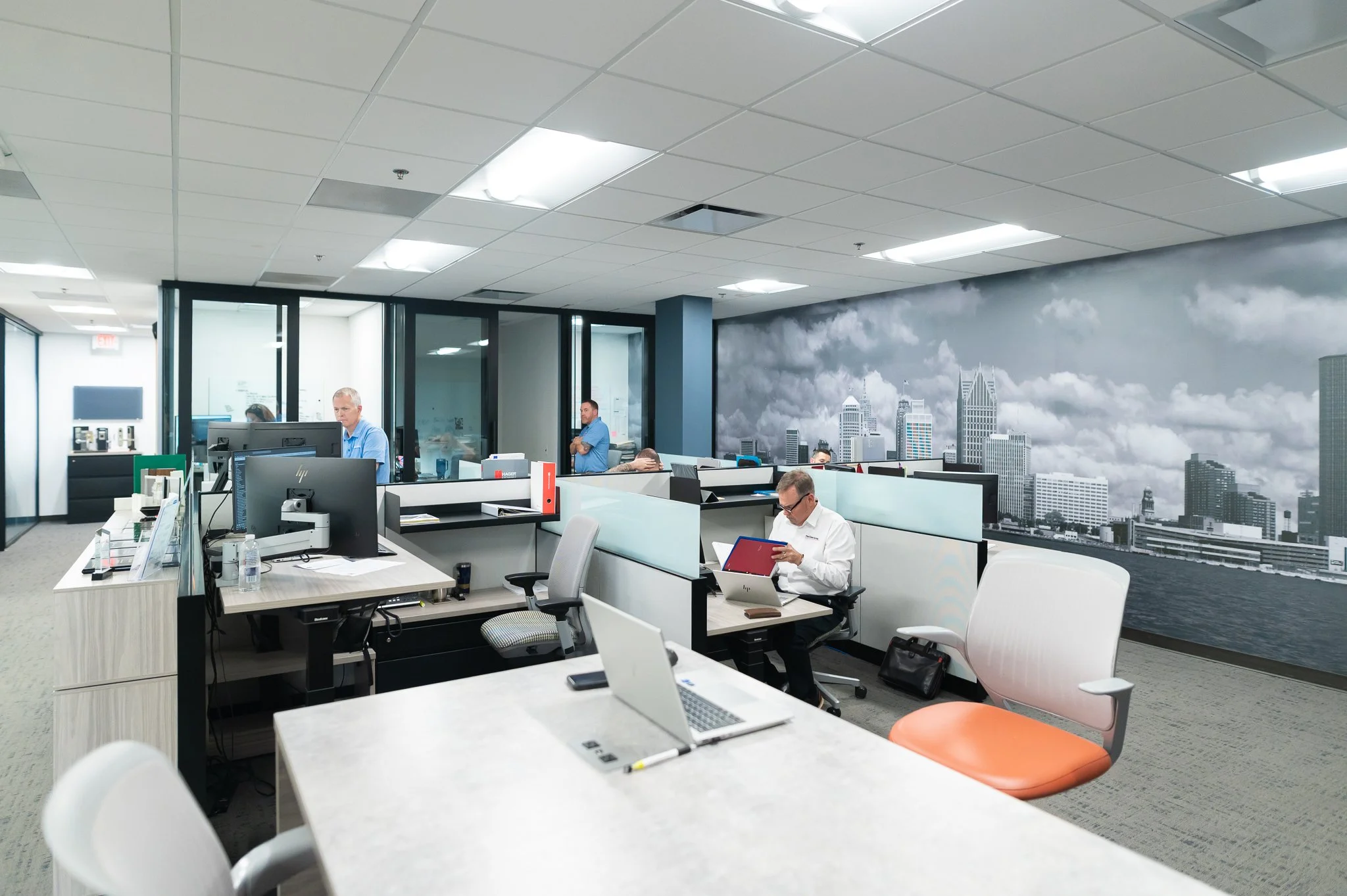 Office with multiple employees working at desks, some using computers, and a cityscape mural on the wall.