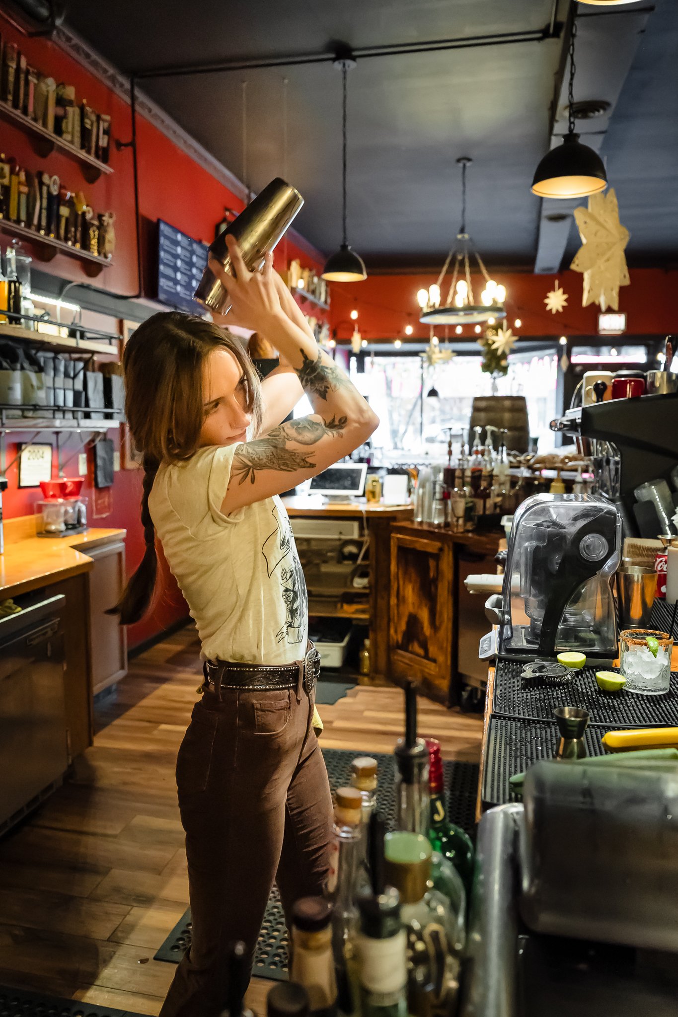A female bartender with tattoos on her arms is preparing a drink behind a bar, holding a shaker above her head. There are bottles and bar tools on the counter, with warm lighting and holiday decorations hanging from the ceiling in the background.