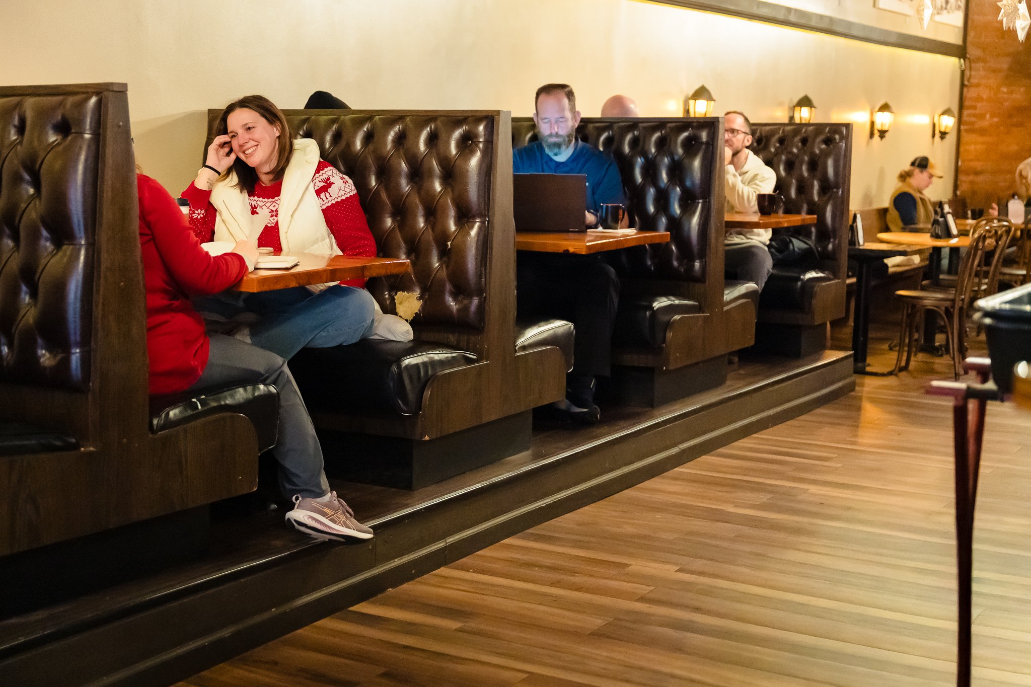 People sitting in restaurant booths, engaged in conversation and working on laptops, with warm lighting and wooden decor.