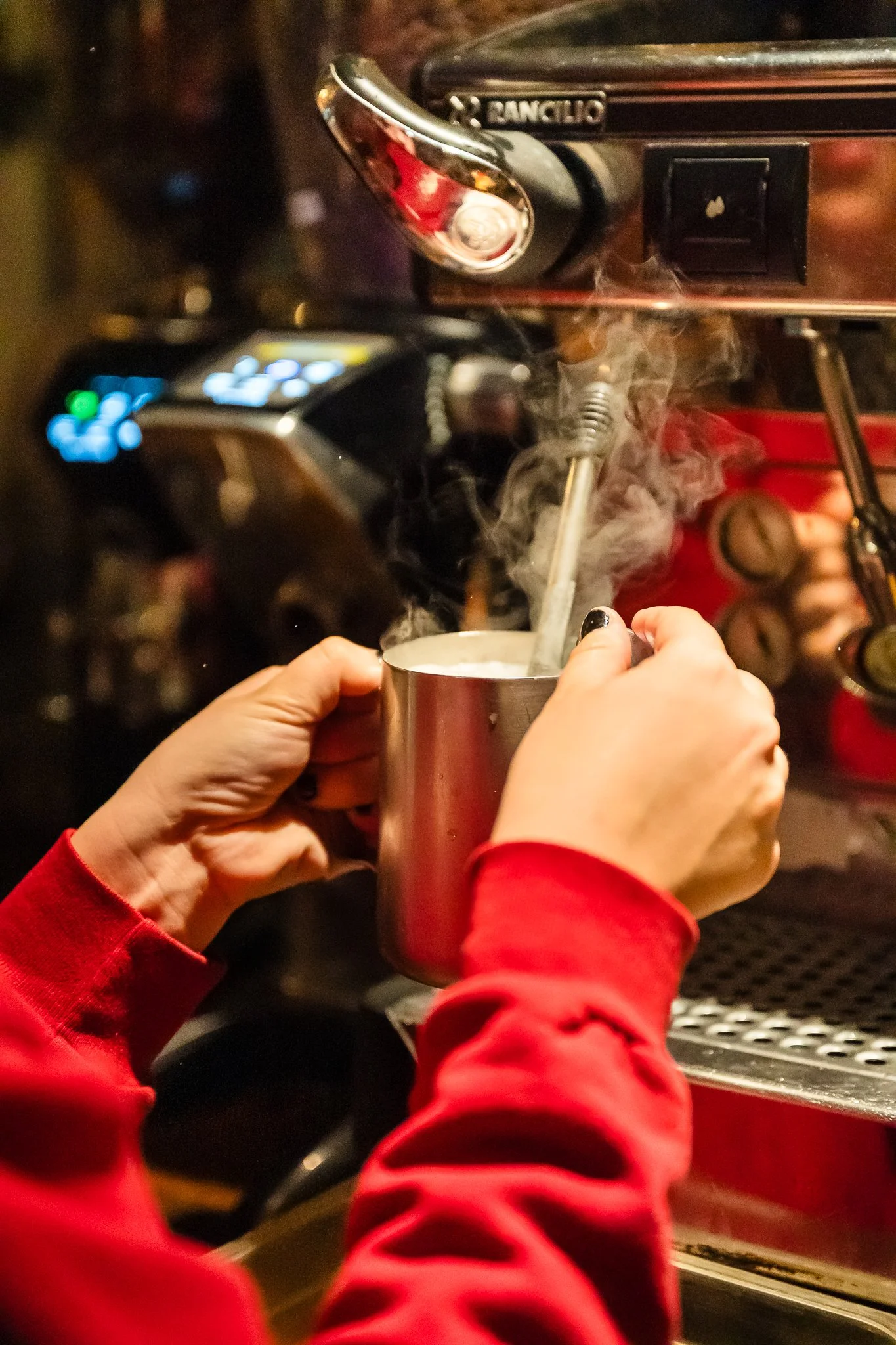 A person in a red shirt holding a insulated mug filled with a hot beverage, steaming, near a commercial espresso machine in a cafe.