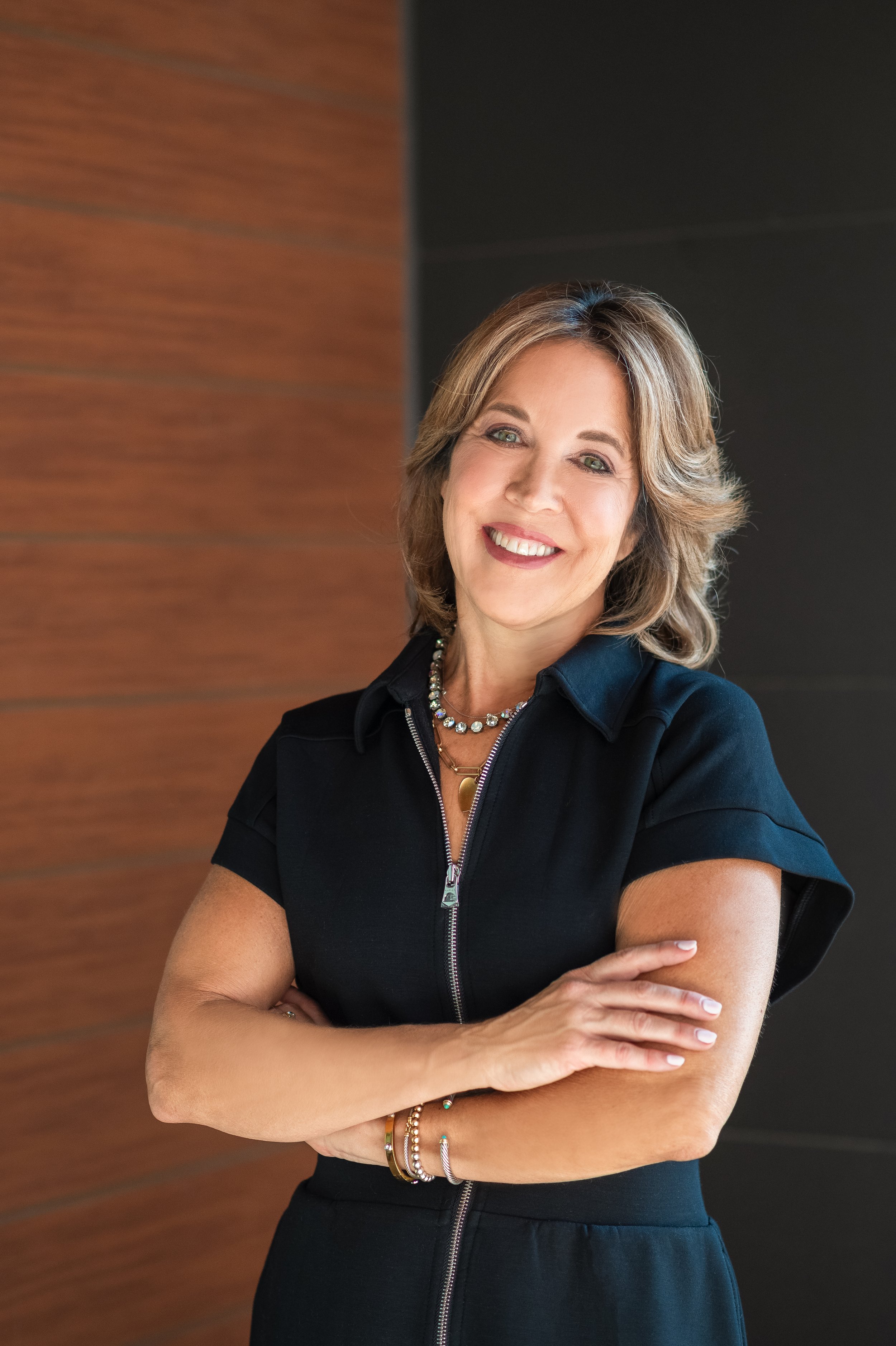 A middle-aged woman with shoulder-length light brown hair, smiling, wearing a black short-sleeve zip-up dress, and jewelry including necklaces and bracelets, standing with her arms crossed against a background of brown and black walls.