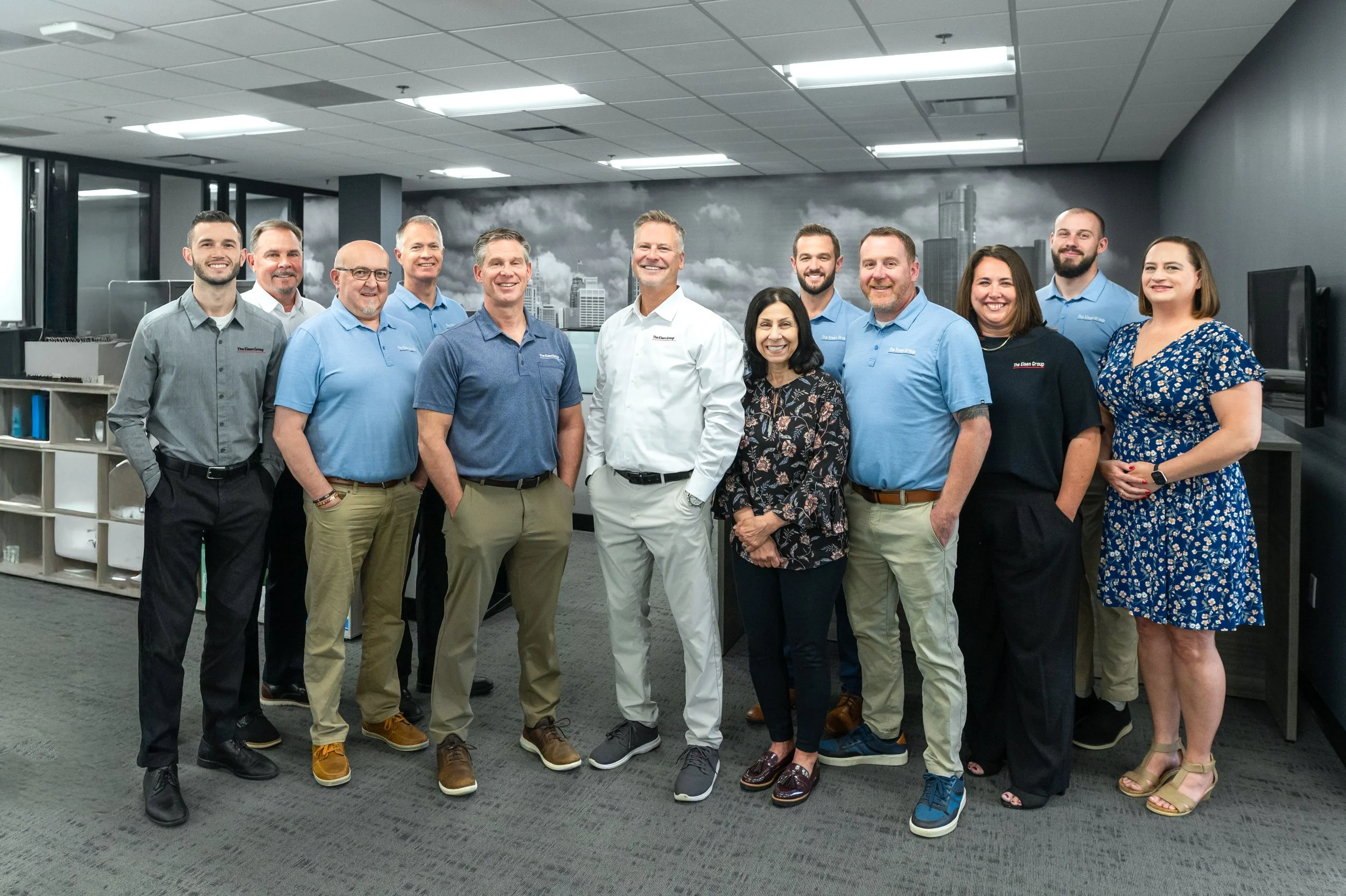 Group of people standing together in an office, smiling for the photo, with a cityscape mural in the background.