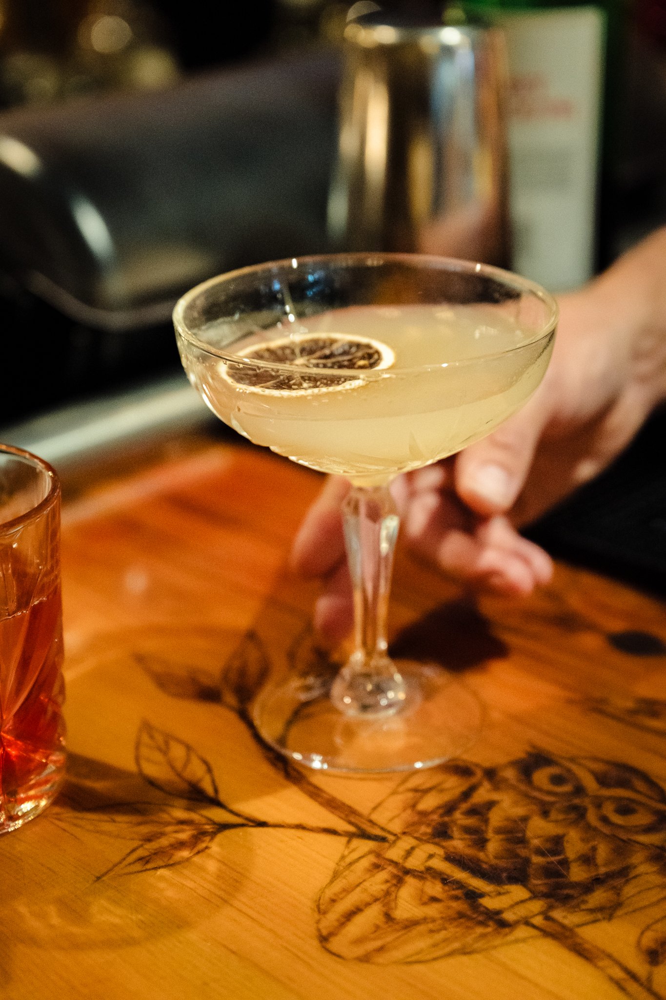 A cocktail in a coupe glass garnished with a dried citrus slice, sitting on a wooden bar counter.