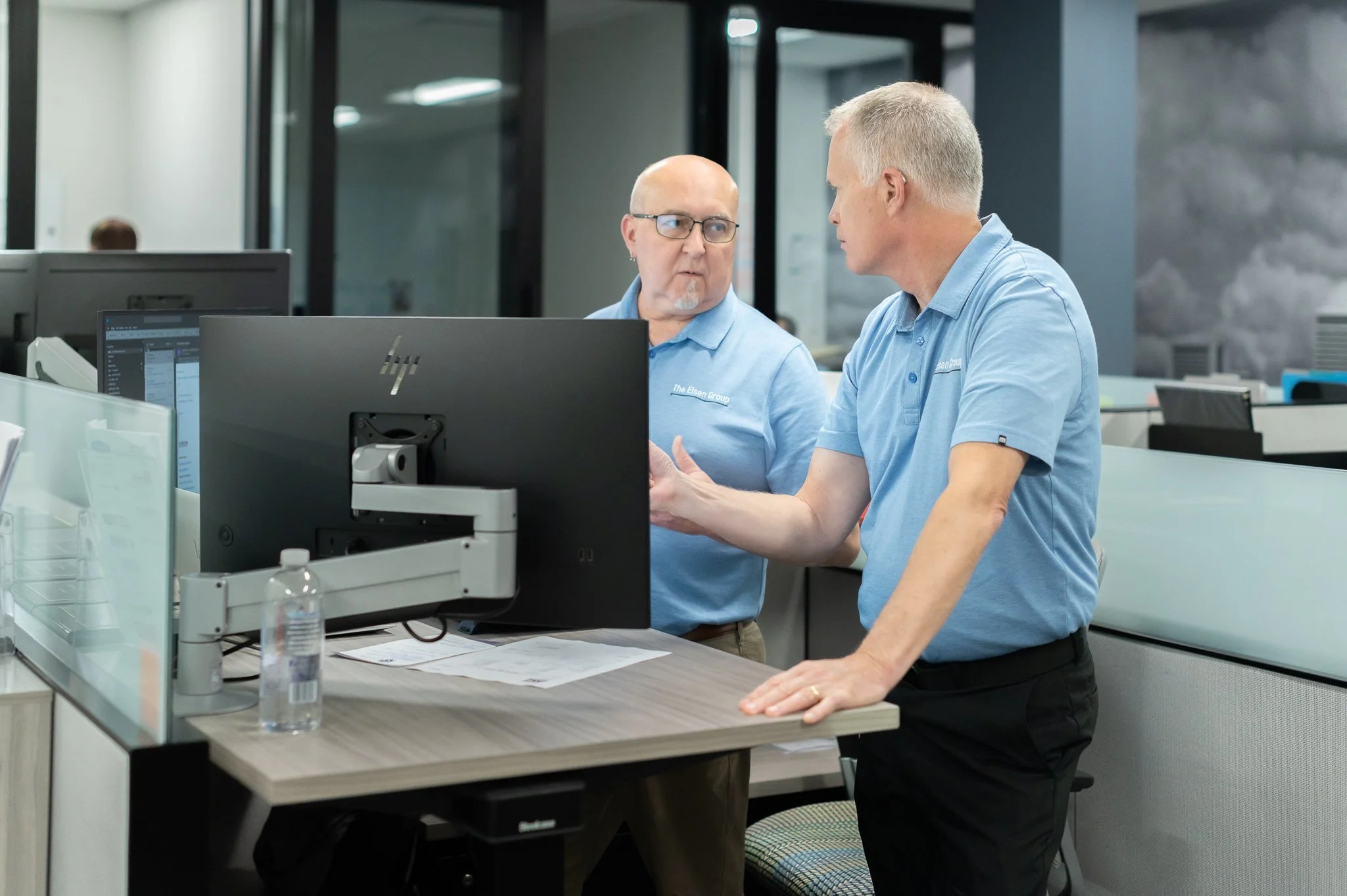 Two men in blue polo shirts having a discussion at a desk in an office, one gesturing towards a computer monitor.