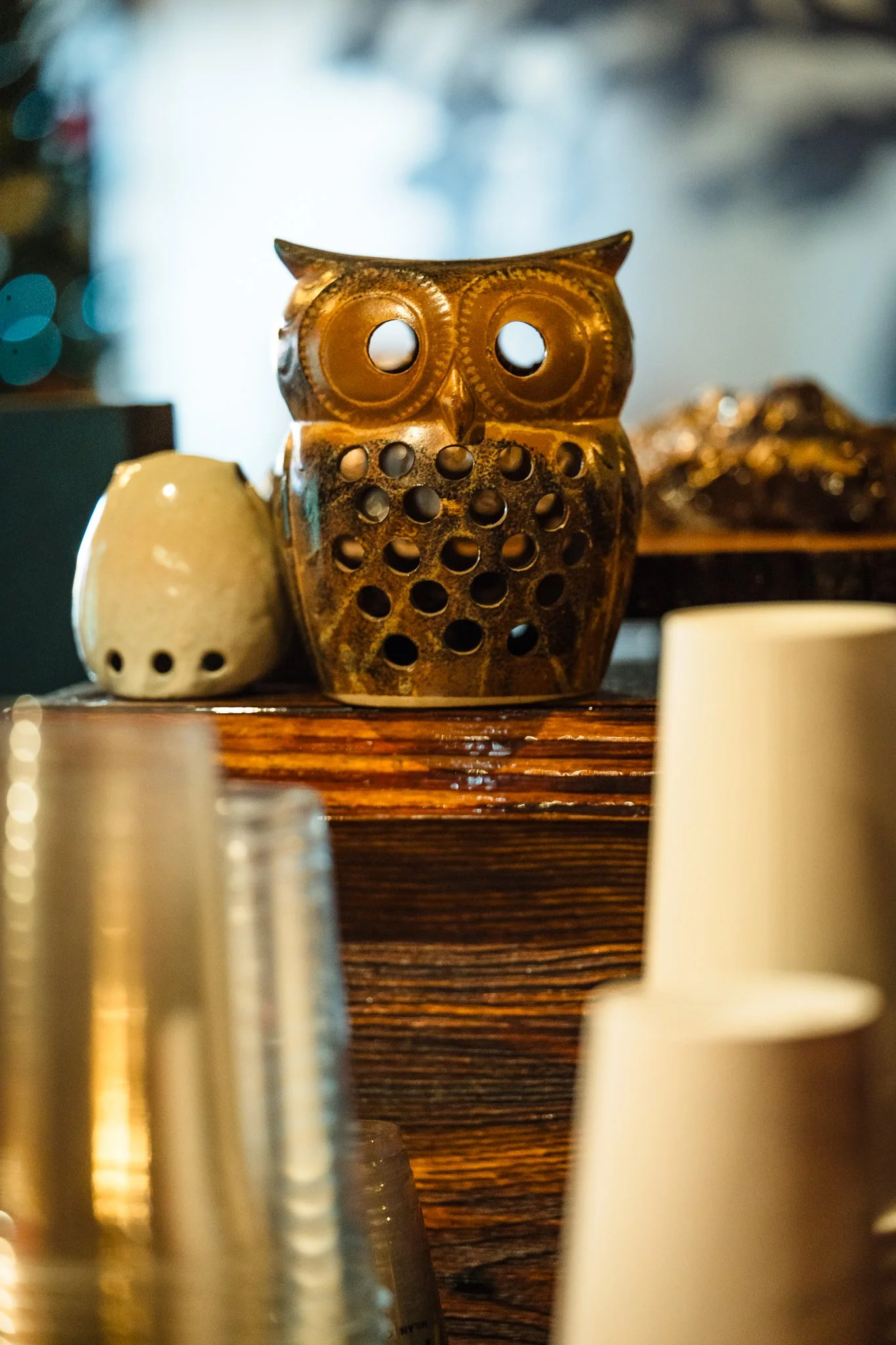 Decorative ceramic owl with circular holes, sitting on a wooden surface, surrounded by stacked cups and other decorative objects.