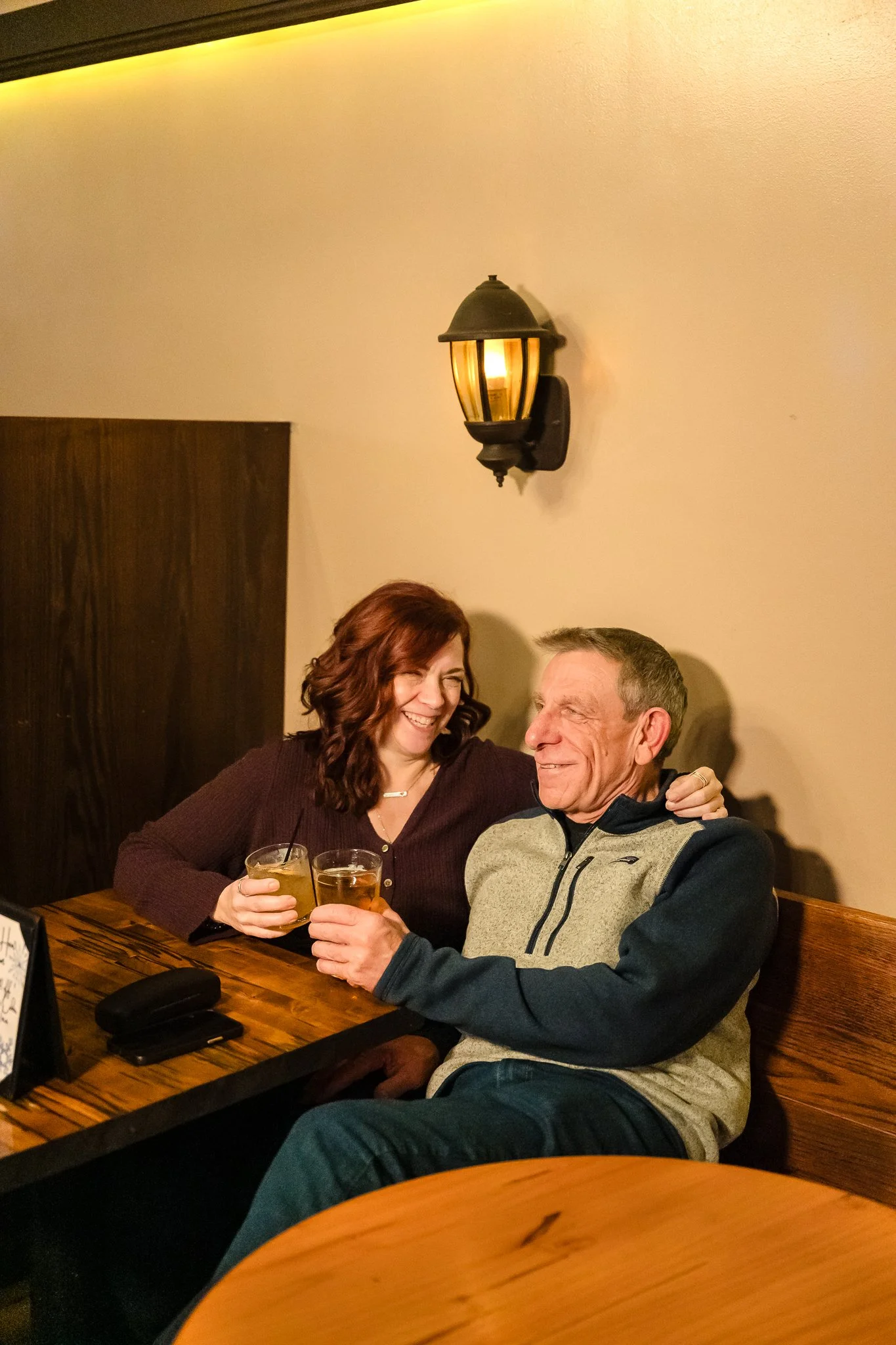 A woman and an older man sitting together at a restaurant table, smiling and holding drinks, with a warm wall-mounted light fixture in the background.