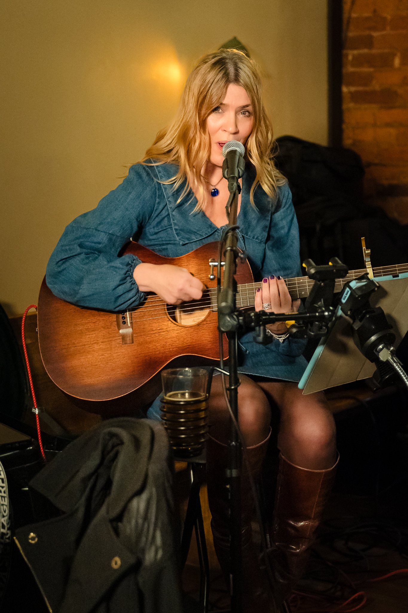 A woman with wavy blonde hair wearing a denim jacket and brown boots is performing live, playing an acoustic guitar while singing into a microphone at a cozy indoor venue.