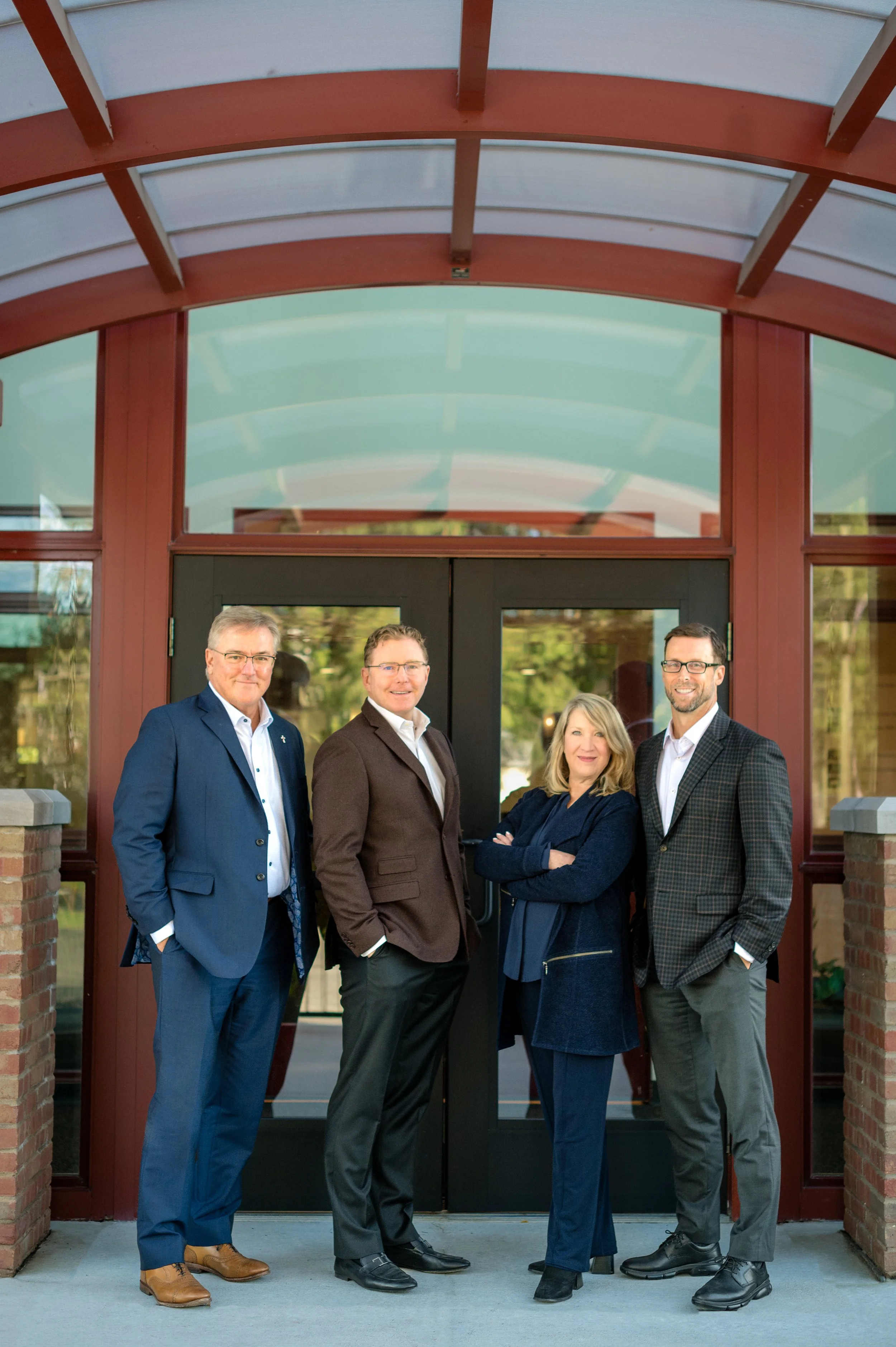 Group of four professionally dressed people standing outside in front of glass doors, with a brick and wood building in the background.