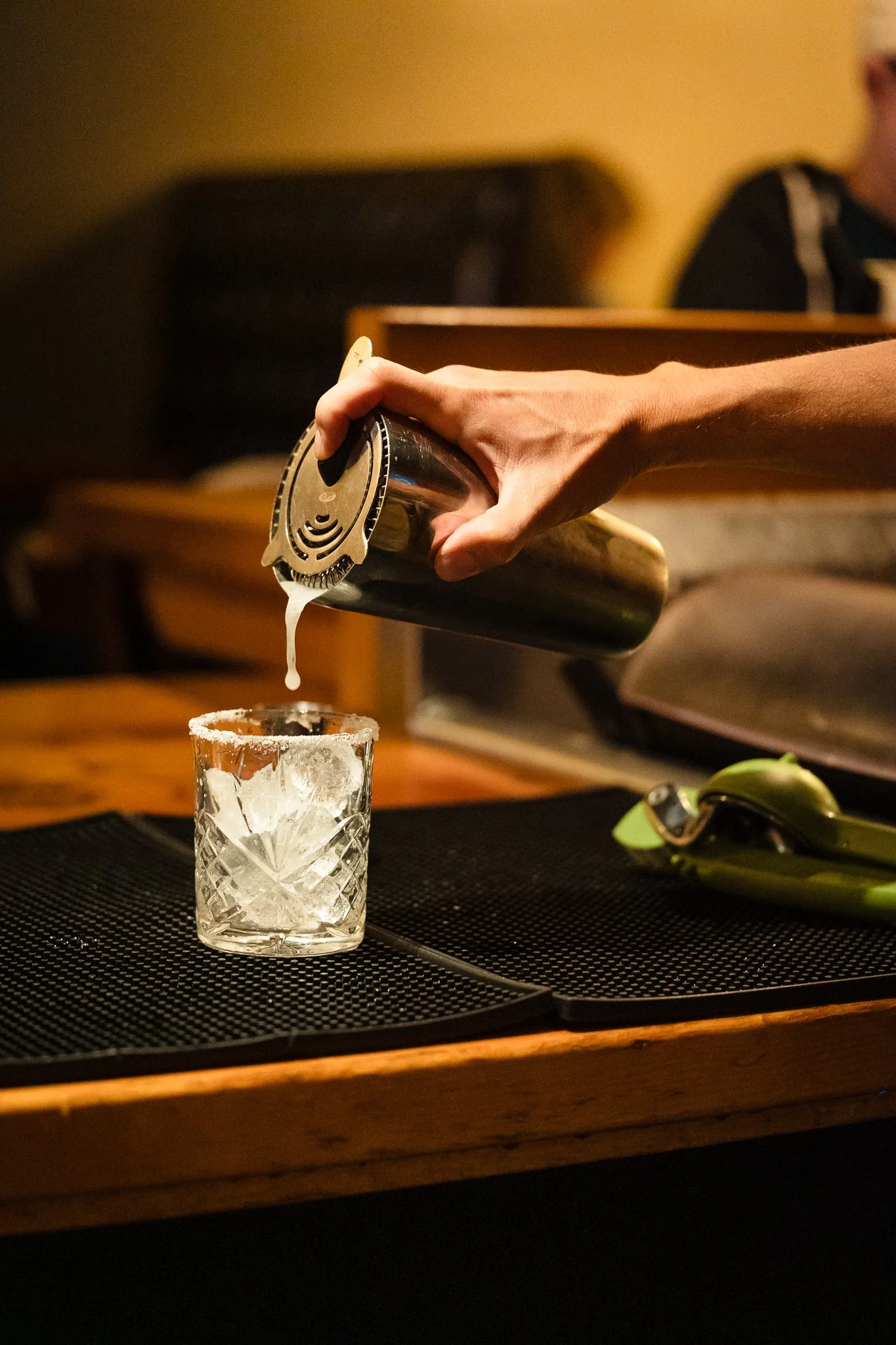 A person pouring a creamy drink into a glass filled with ice on a wooden bar counter.