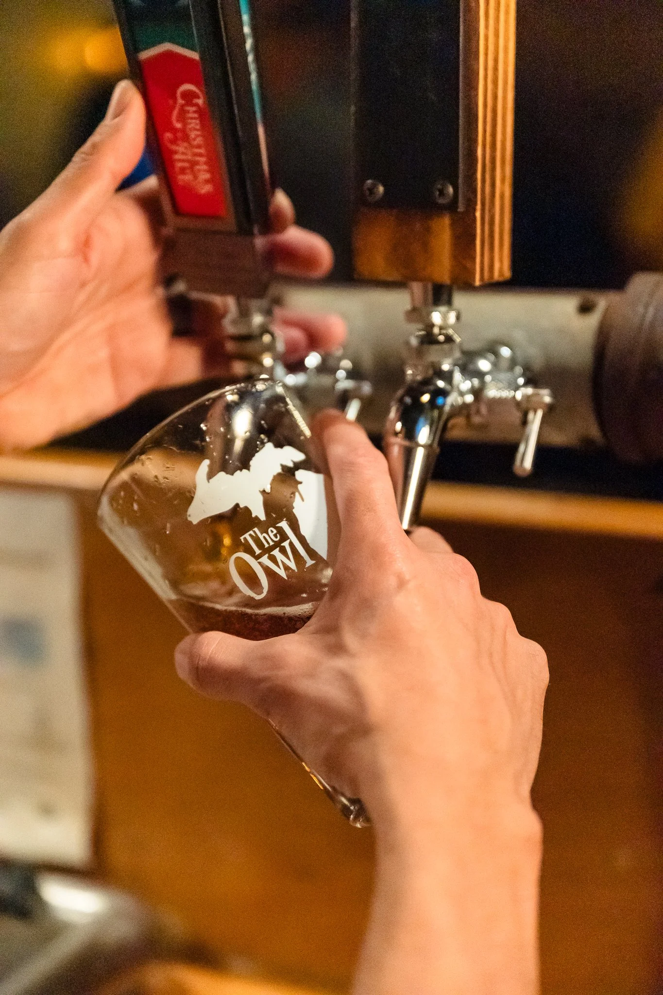 Person pouring draft beer into a glass with 'The Owl' logo in a bar or brewery setting.