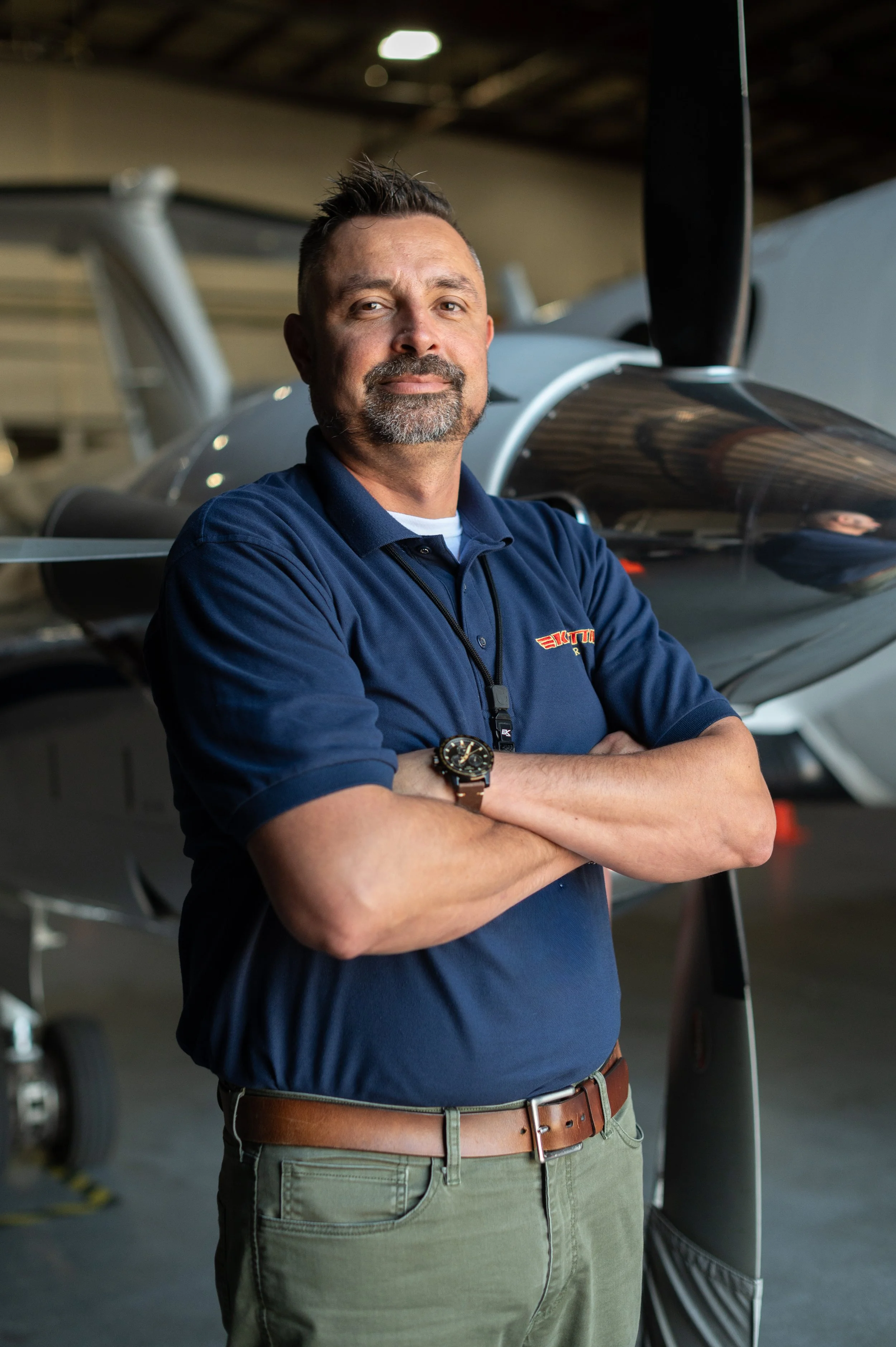 A man with a beard and short hair standing with arms crossed in front of a small airplane inside a hangar.
