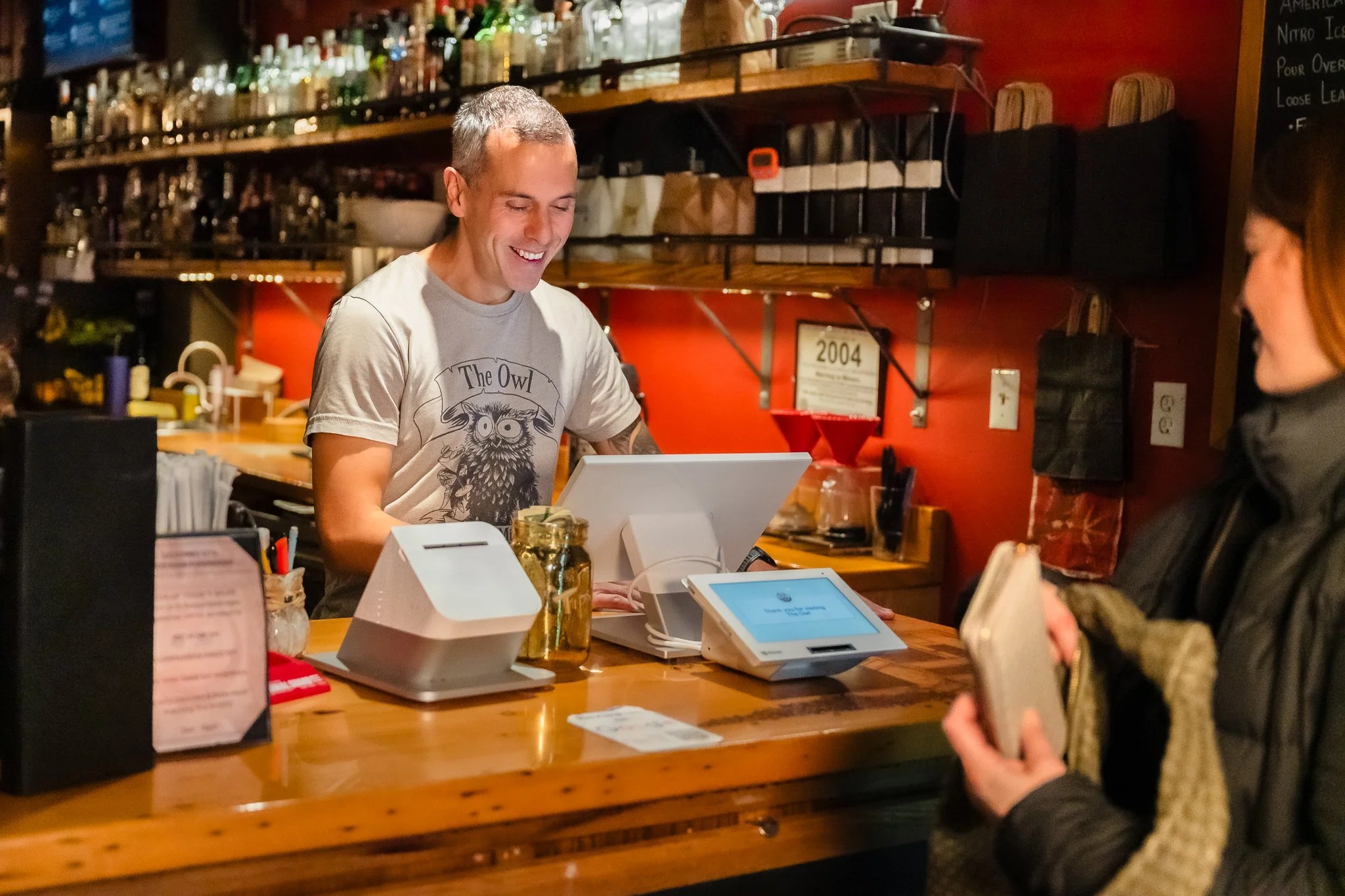 A man smiling while working behind a bar counter, with a woman holding a smartphone in front of him, inside a dimly lit bar or restaurant.