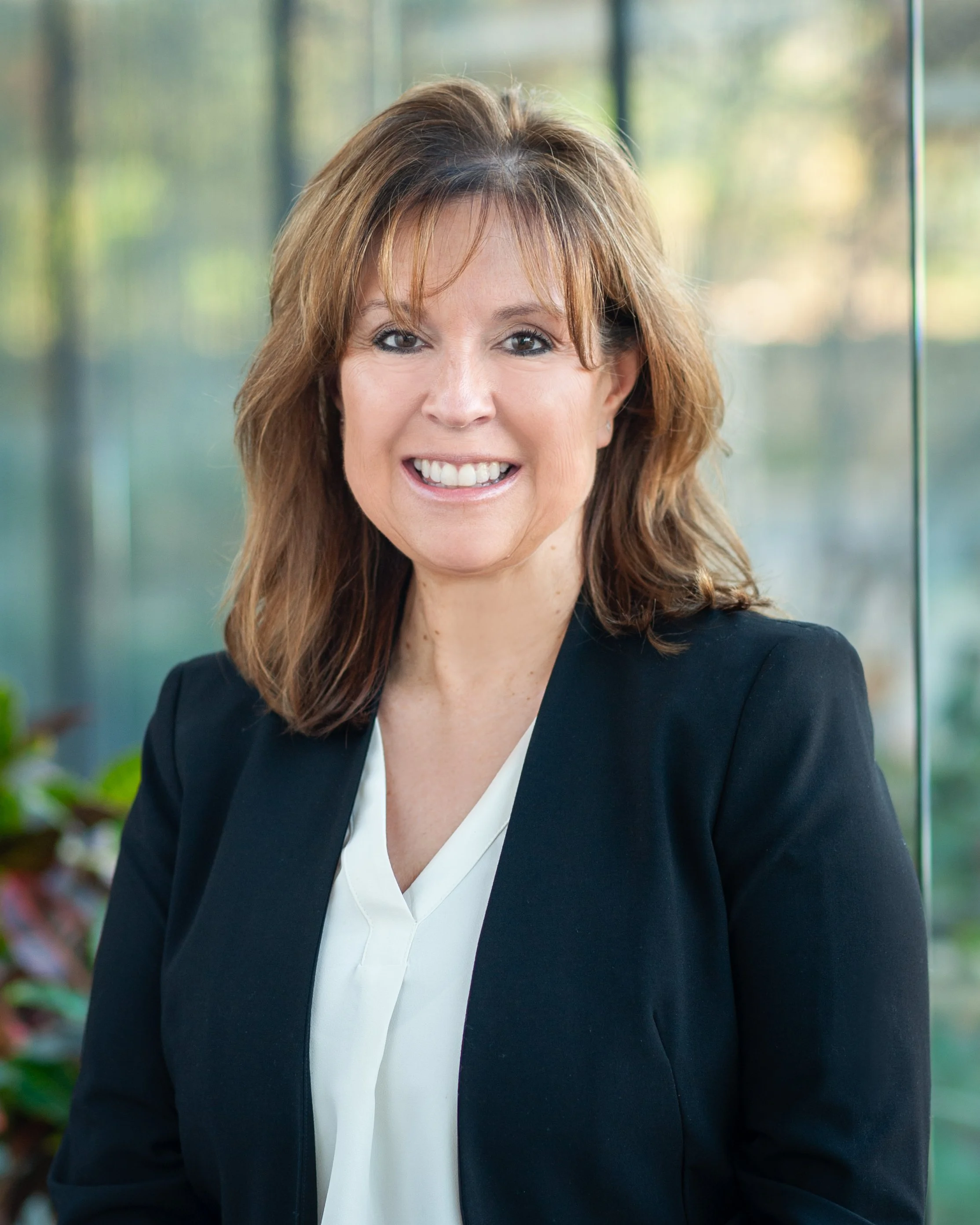 A woman with shoulder-length brown hair smiling, wearing a white blouse and a black blazer, standing in front of a window with greenery outside.