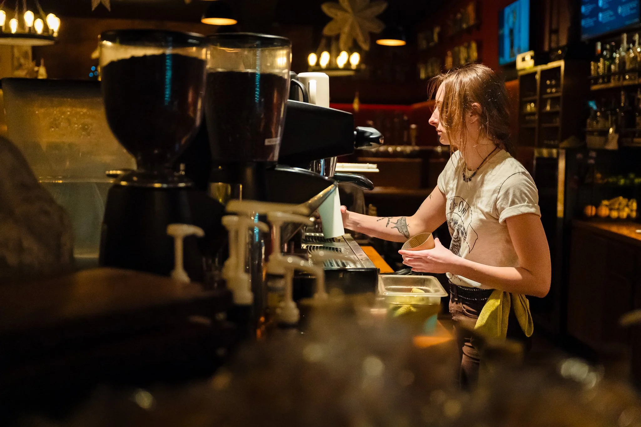A young woman with tattoos and a graphic t-shirt working as a barista behind a coffee counter in a dimly lit cafe or bar, preparing a drink. There are two large glasses of dark beer in the foreground.