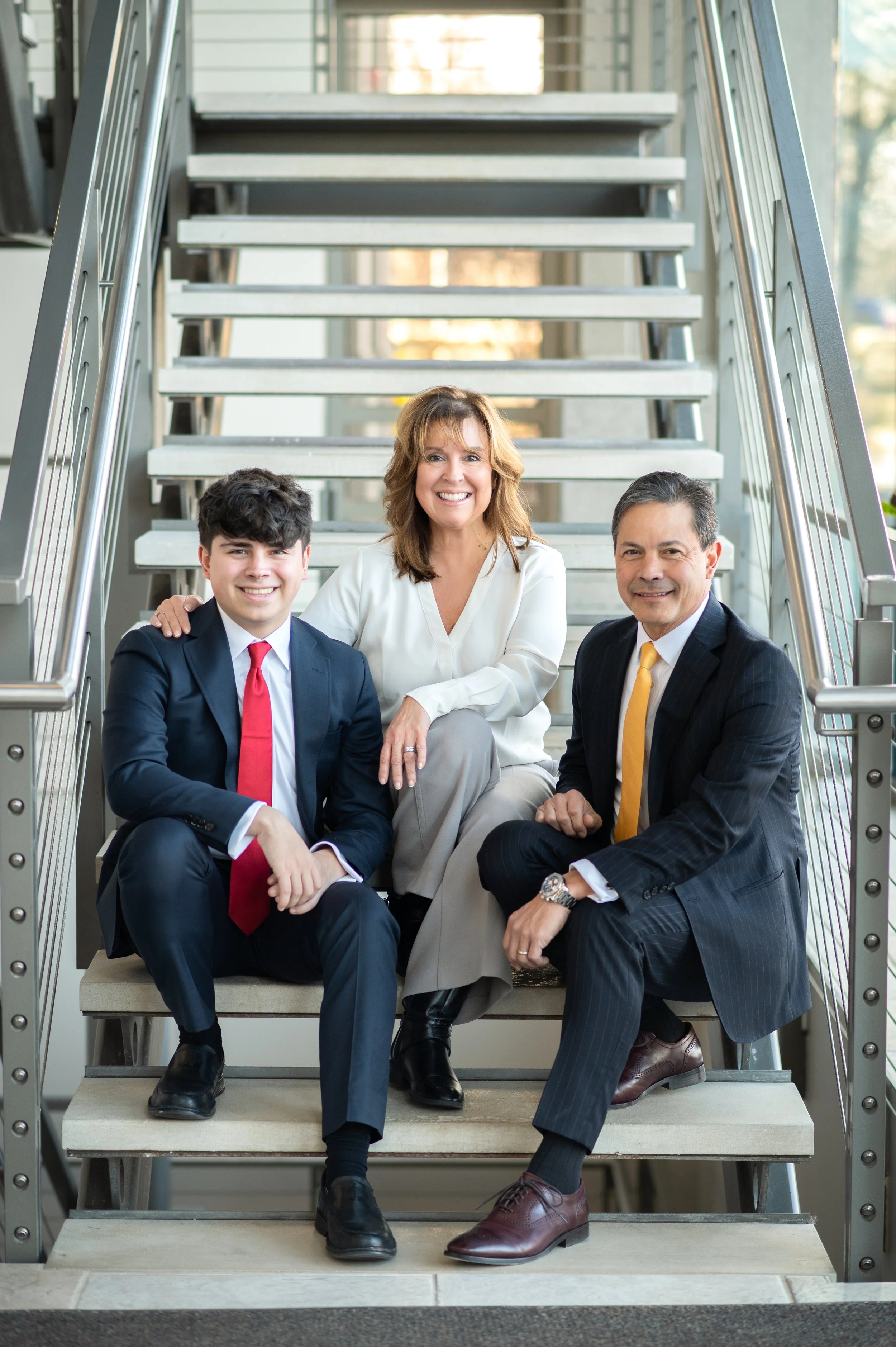 A woman and two men sitting on stairs, smiling at the camera. The woman has shoulder-length brown hair and is wearing a white blouse. The young man on the left has dark hair, a navy suit, and a red tie. The man on the right has dark hair, a dark pins