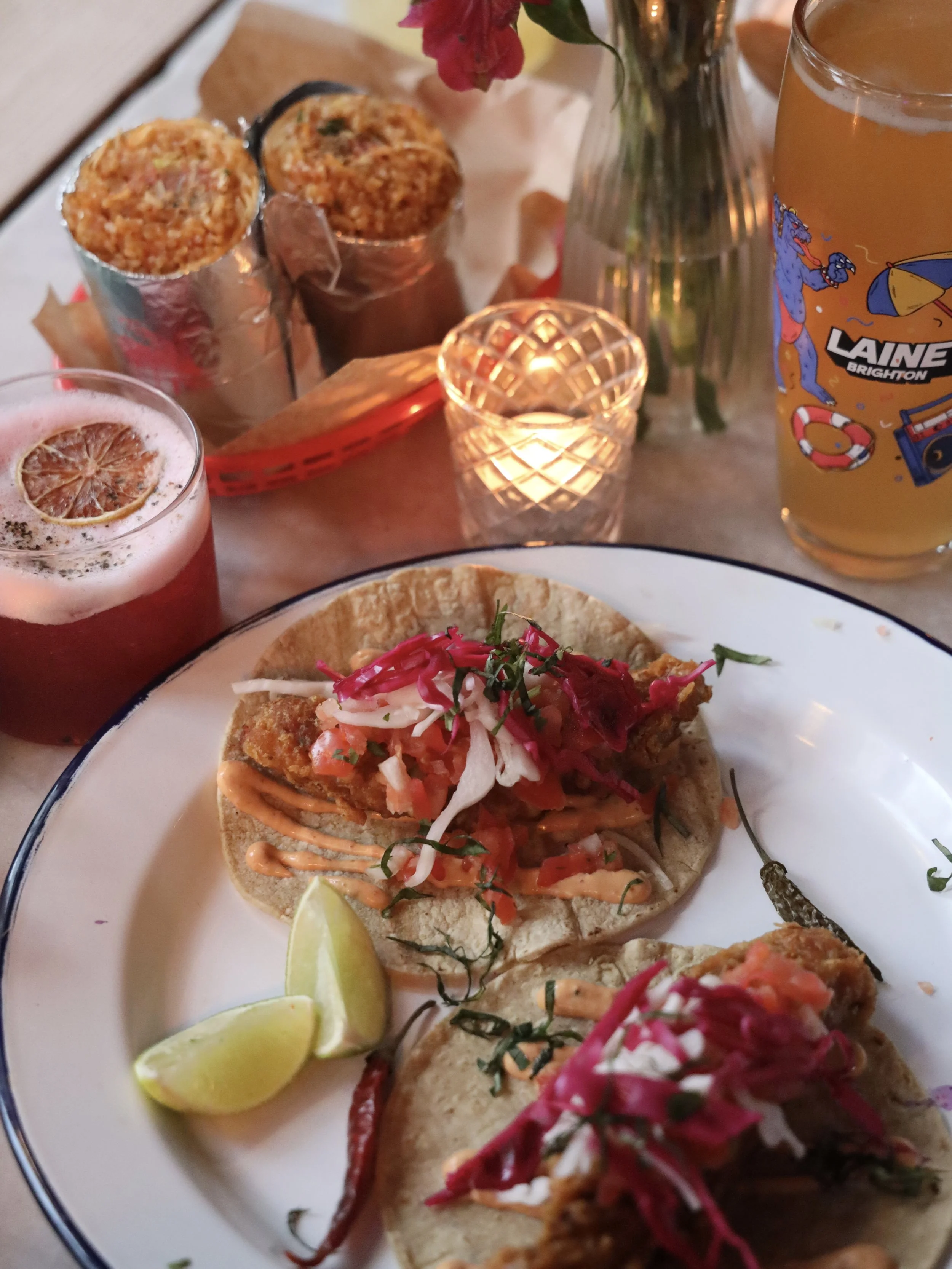 A plate of fish tacos garnished with chopped vegetables and herbs, served with lime wedges. In the background, two cans of what appears to be canned or bottled drinks, a glass of beer, a floral arrangement, and a lit candle are visible on the table.