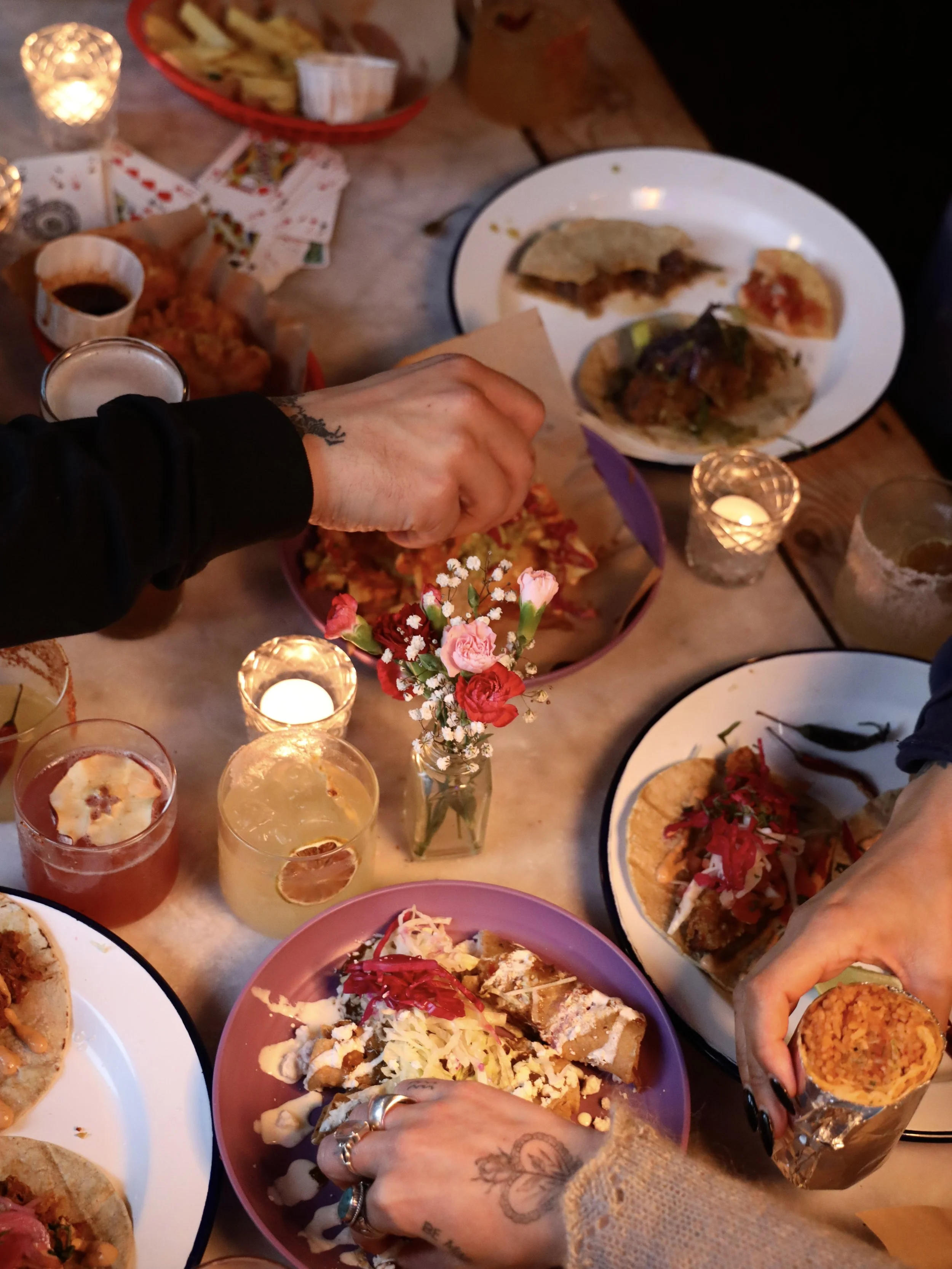 A table with several plates of food including tacos, nachos, and other Mexican dishes, along with drinks, flowers, and candles. People are reaching for the food.