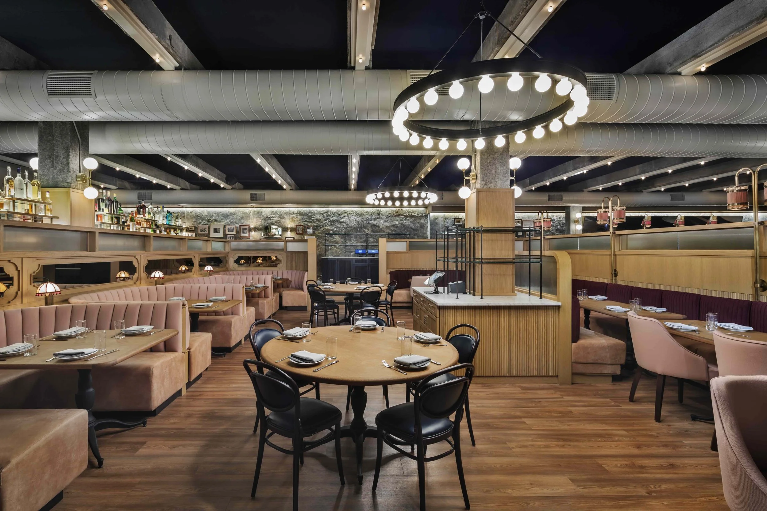 Contemporary restaurant interior with blush banquette seating, exposed ductwork, circular pendant lighting, wood floors, and set dining tables.