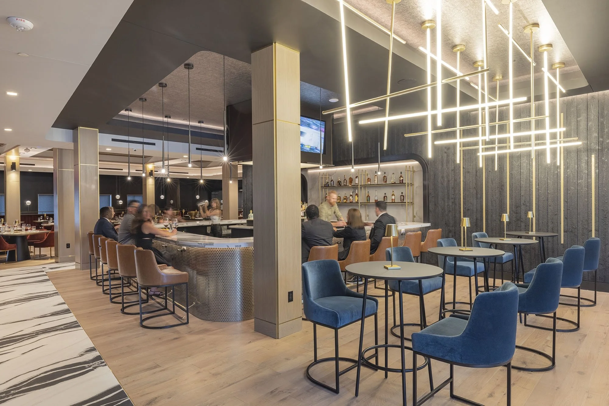 Contemporary bar and lounge with dark finishes, sculptural linear lighting, blue seating, wood floors, and guests gathered at the bar.
