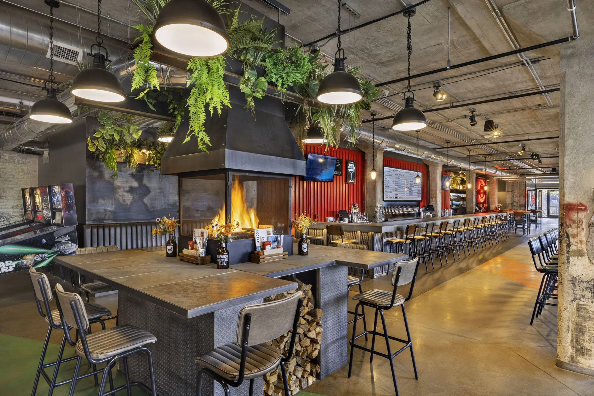 Industrial-style restaurant interior with exposed concrete, hanging greenery, a fireplace, black pendant lights, long bar, and metal-framed tables.