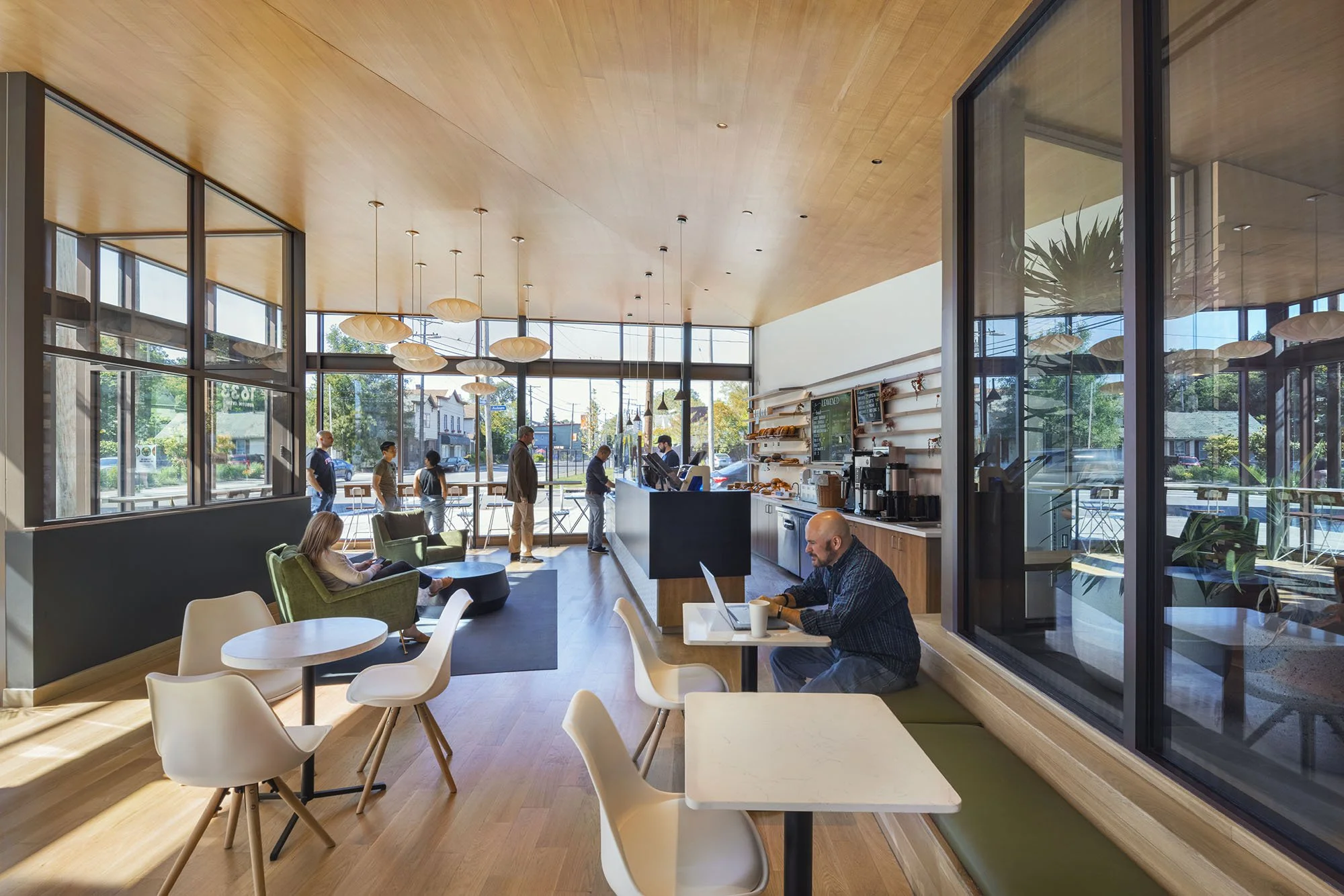 Bright restaurant and café interior with floor-to-ceiling windows, wood ceiling, modern seating, and guests using the dining and work areas.