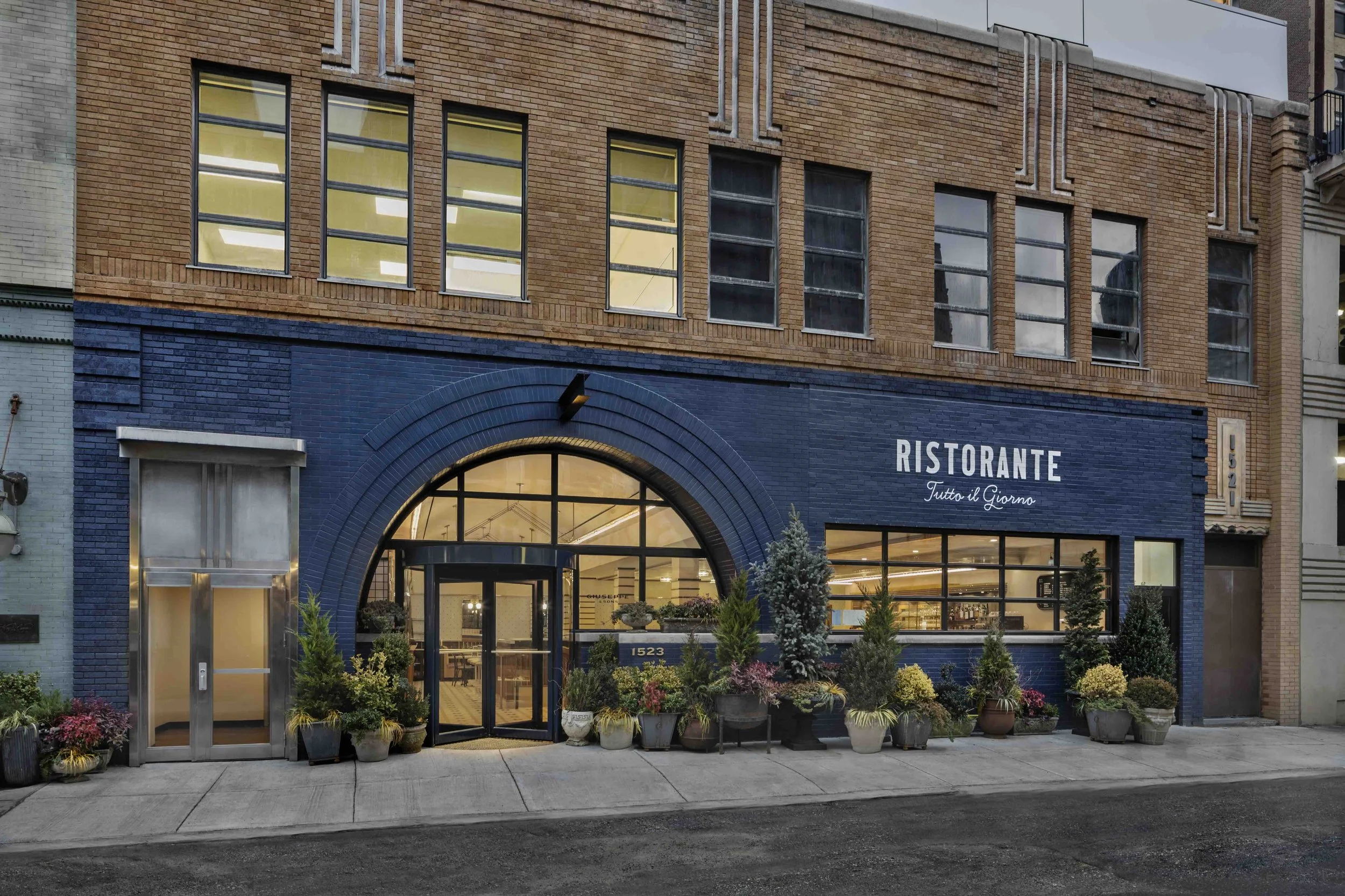 Street-level restaurant exterior with dark blue brick facade, arched entrance, large windows, landscaping, and illuminated interior seating.