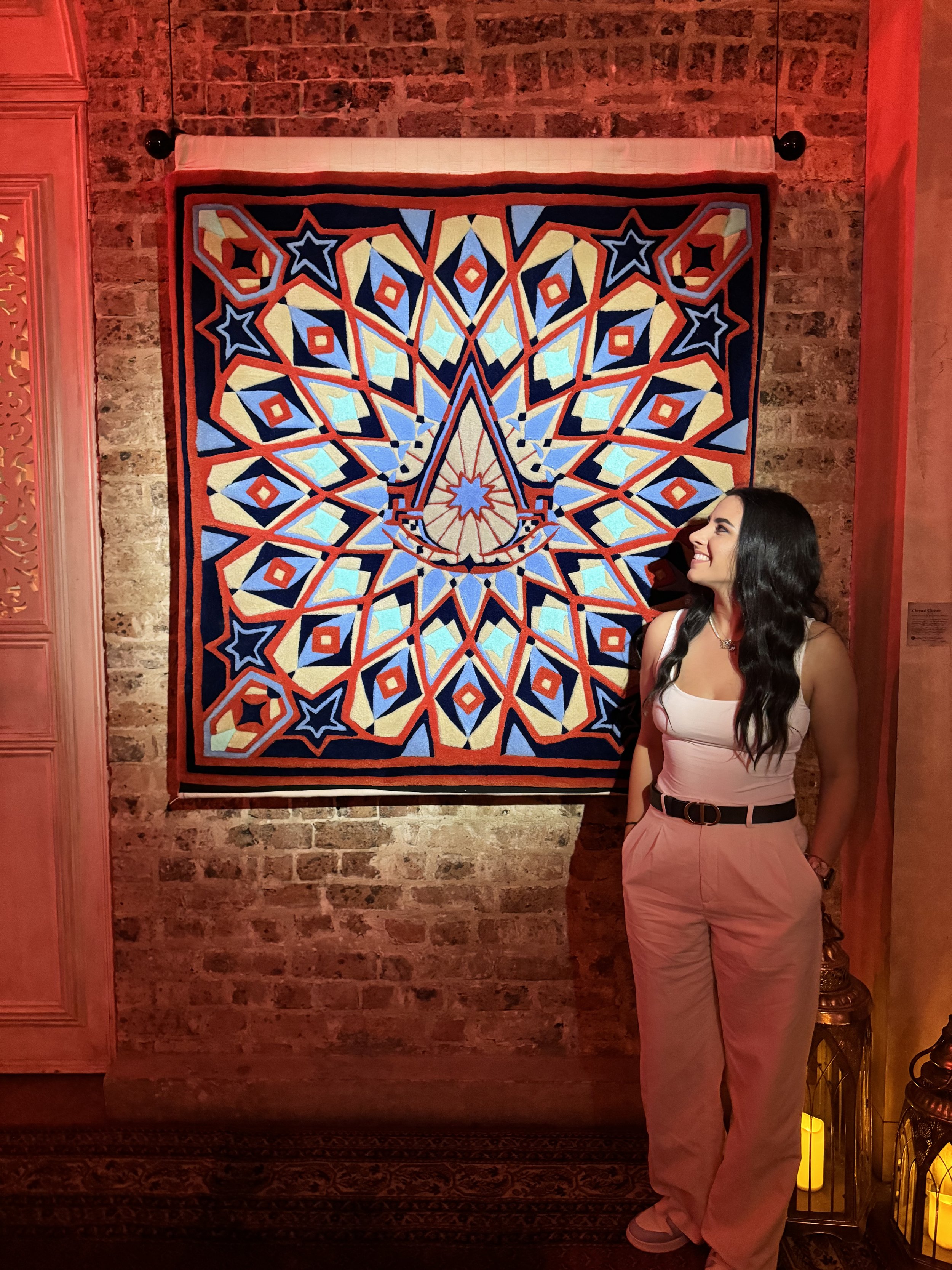 A woman stands smiling with her hands in her pockets, next to a colorful geometric textile hanging on an exposed brick wall, with lanterns on the floor nearby.
