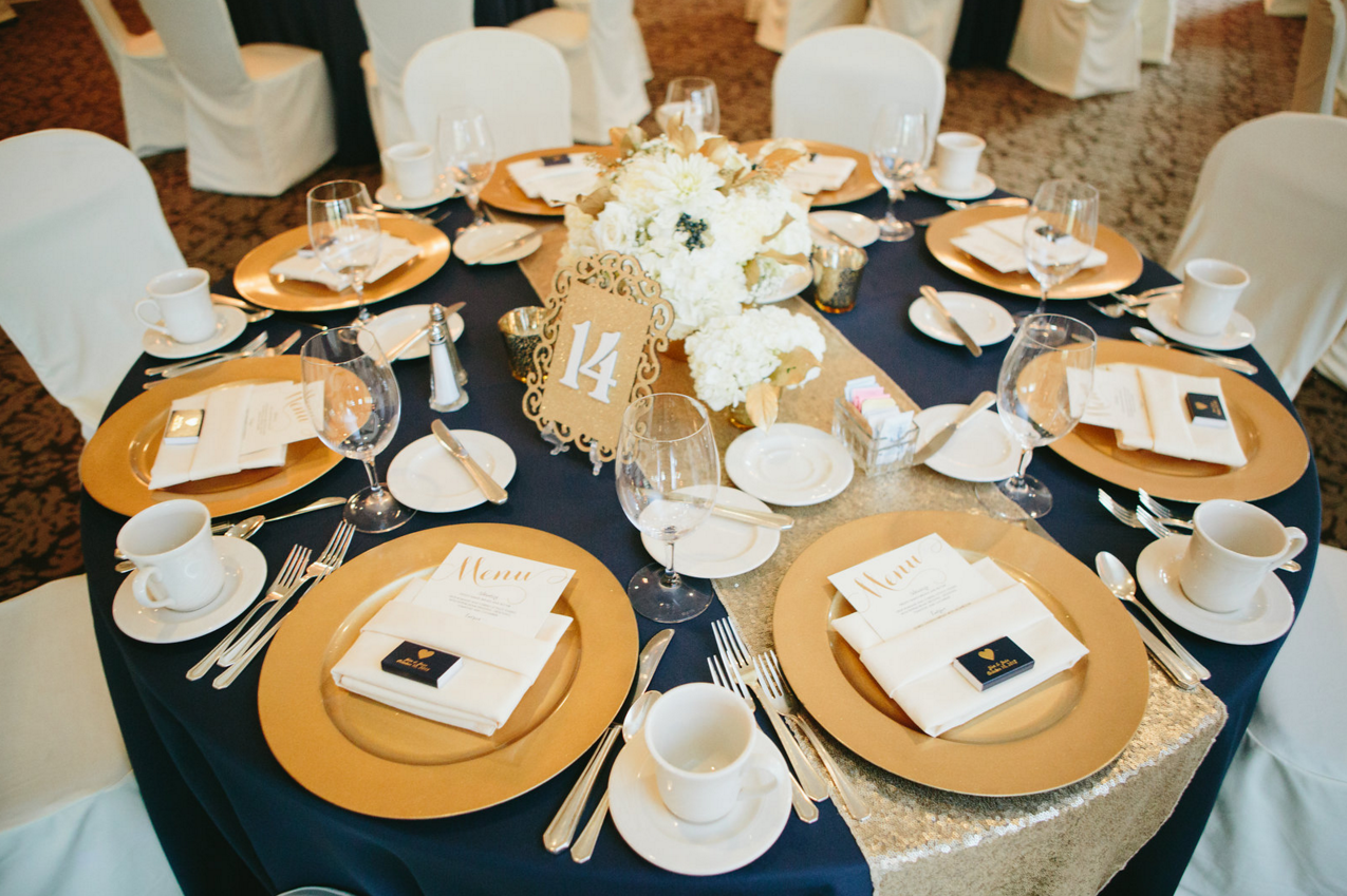 gold sequin table runner on a wedding reception table with gold table numbers, gold chargers and gold candle votives
