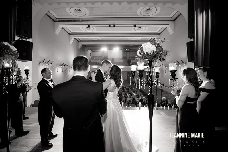 The first kiss of husband and wife in black and white shot from behind towards guests overseeing tall candleabras with crystals and flowers and candles