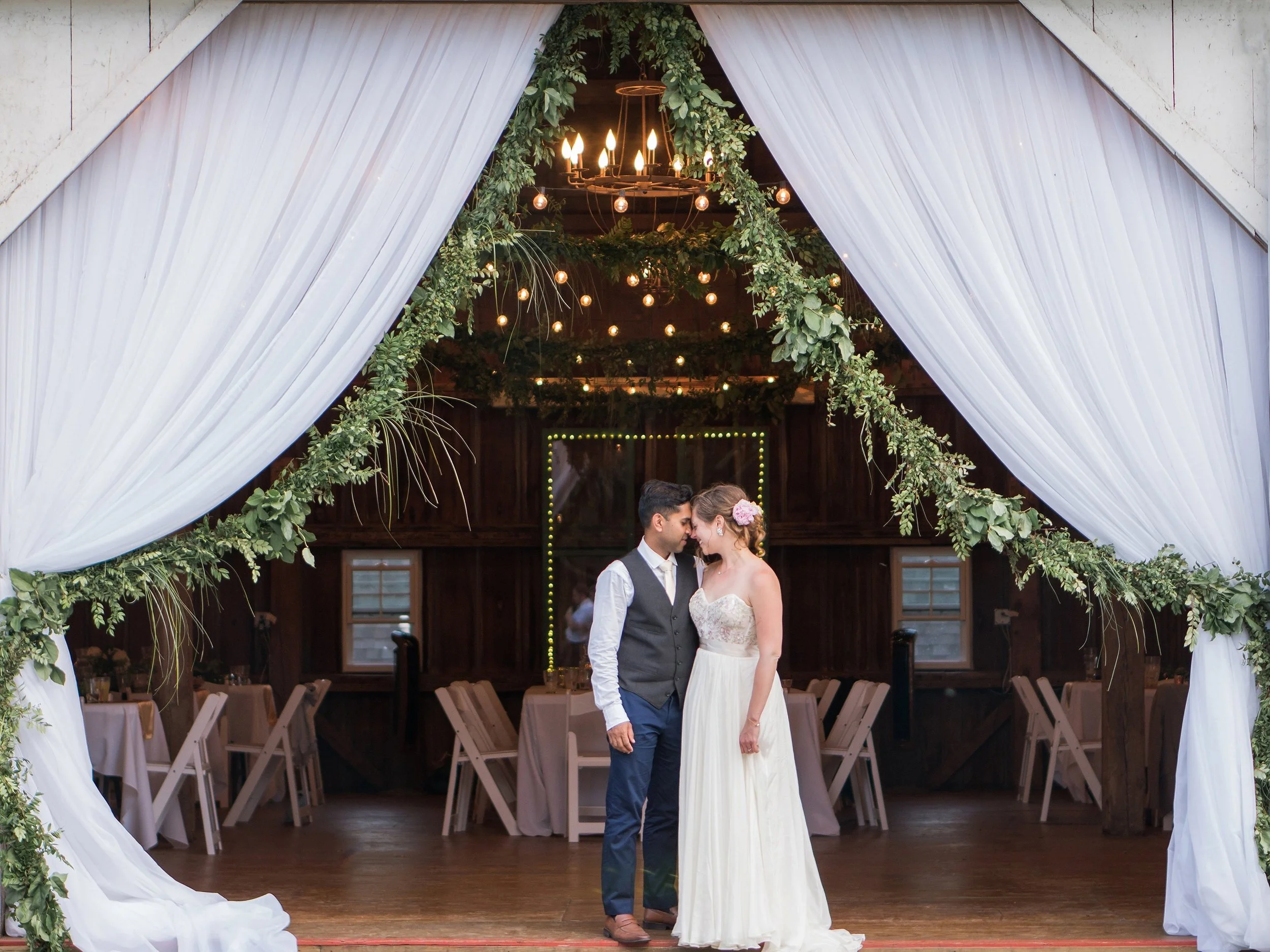 Newlywed couple in a barn entrance draped with white curtains pulled back with hanging greenery
