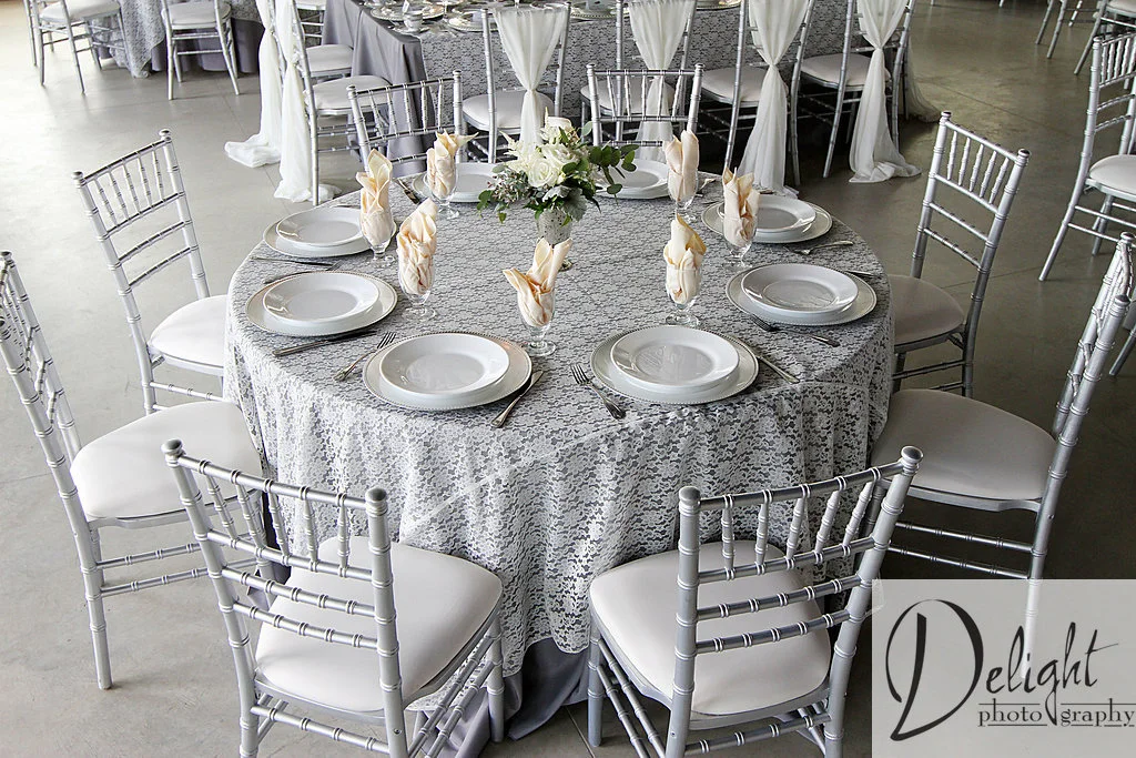 reception table styled with grey linens and lace overlays. The ivory napkins placed in glasses and a floral arrangement for a centerpiece