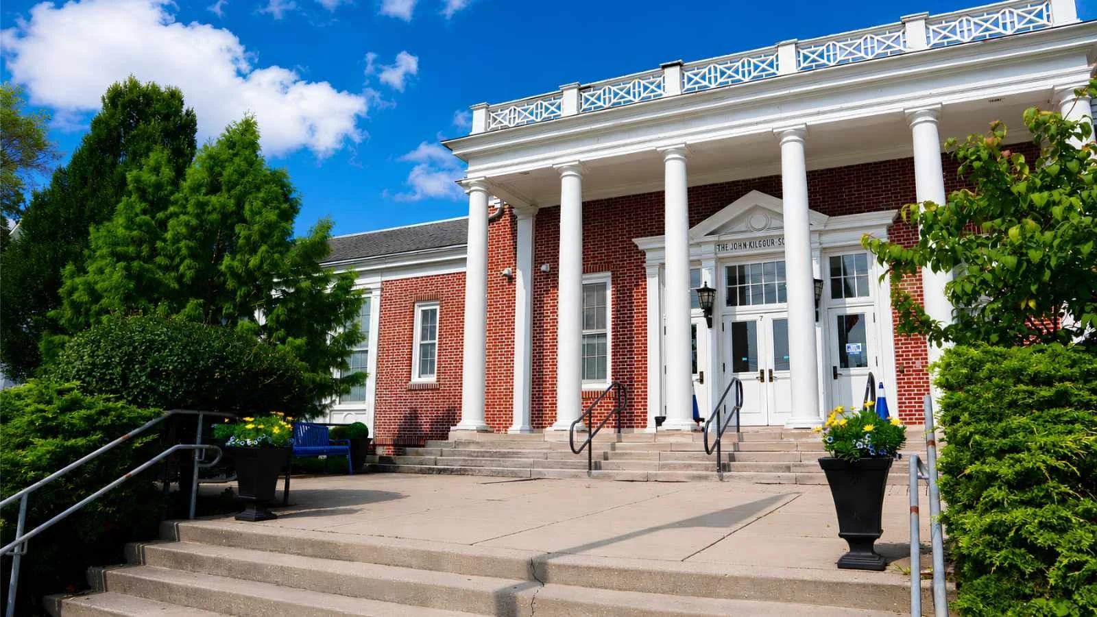 Front entrance of a brick building with white columns, stairs, green bushes, potted flowers, and a blue bench under a partly cloudy sky.