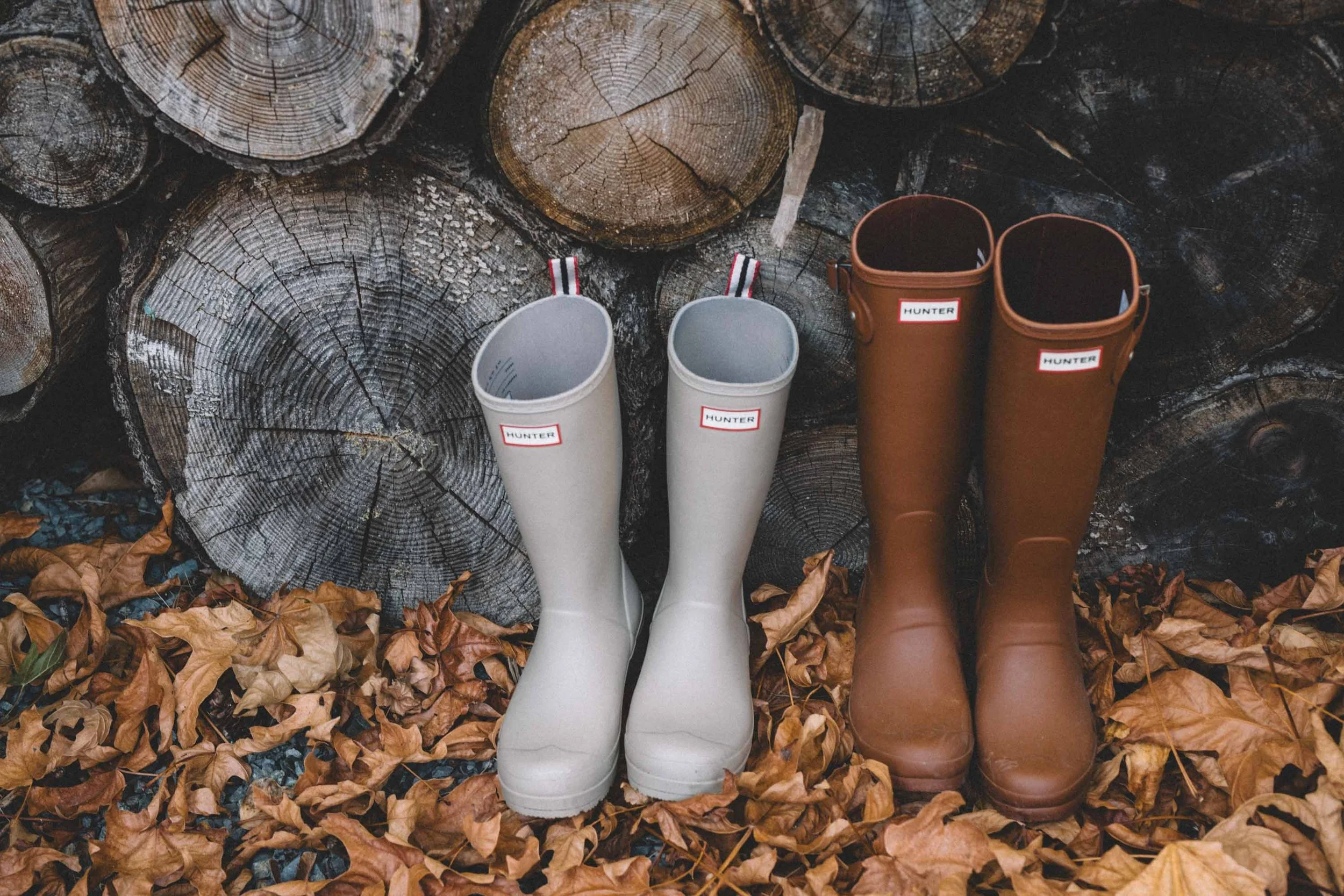 Four pairs of Hunter rain boots arranged in front of a stack of cut logs, with fallen leaves on the ground.