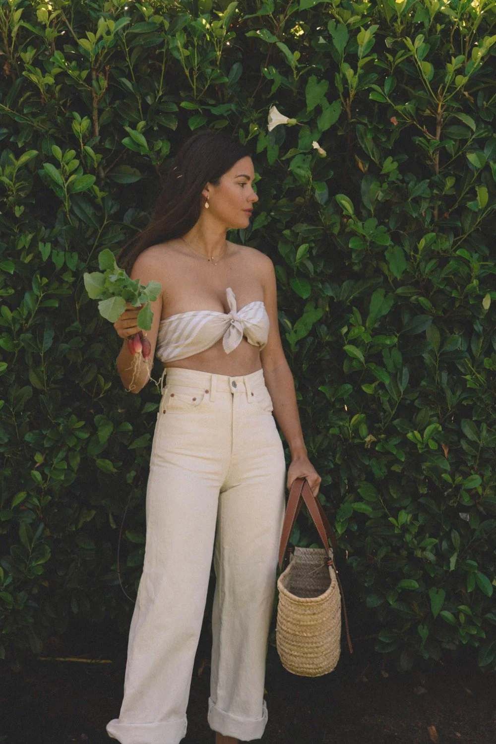Woman holding fresh radishes in a garden wearing a neutral summer gardening outfit with a bandeau top and high-waisted pants