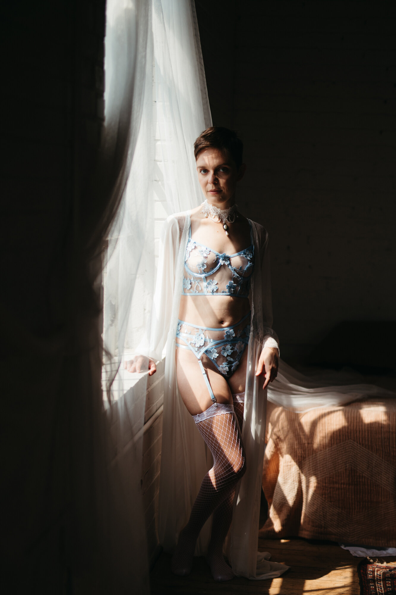 Genderqueer person in victorian style blue lingerie standing by a window