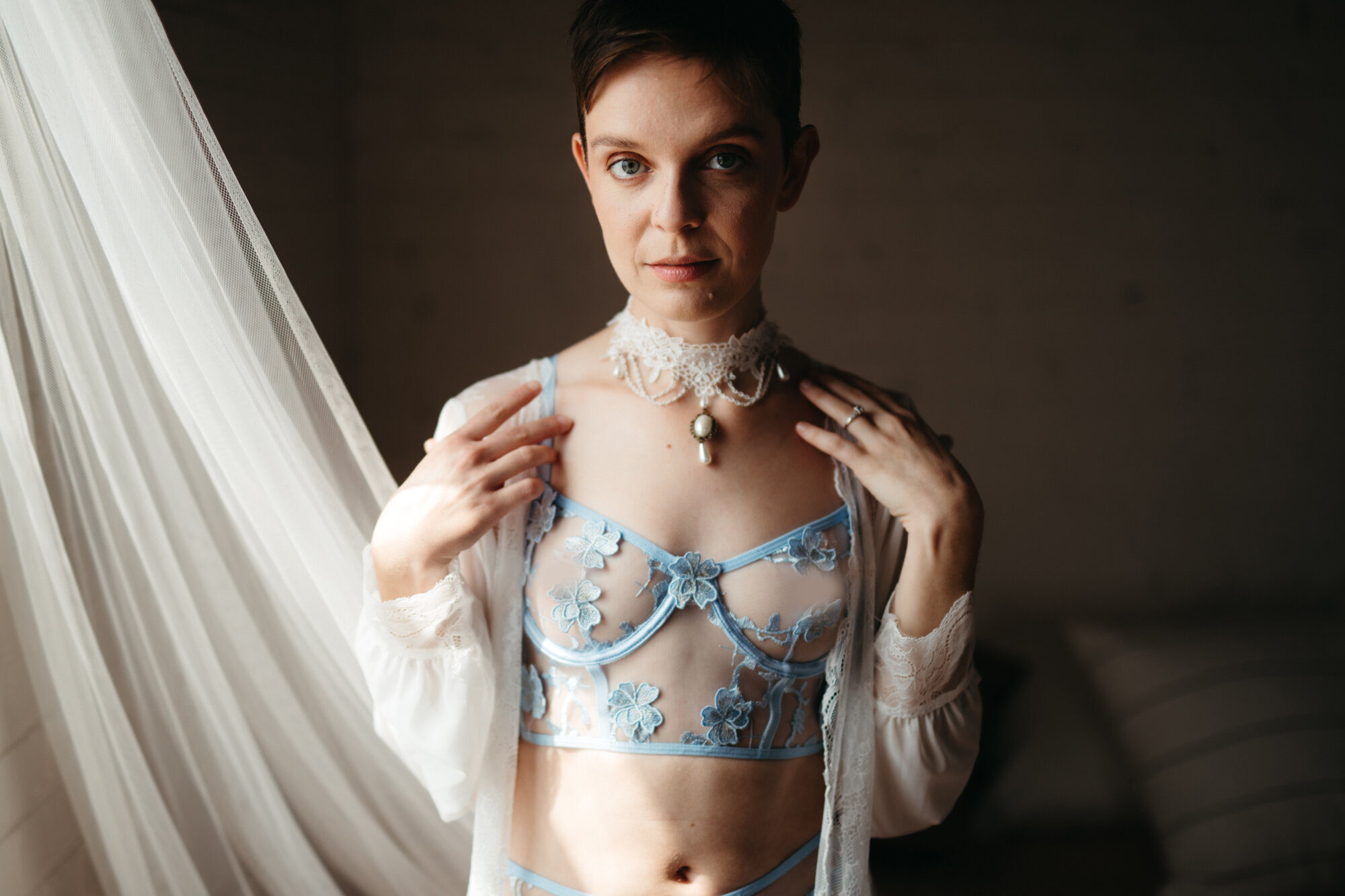Genderqueer person wearing blue lingerie set and a white robe while lightly touching their collarbone
