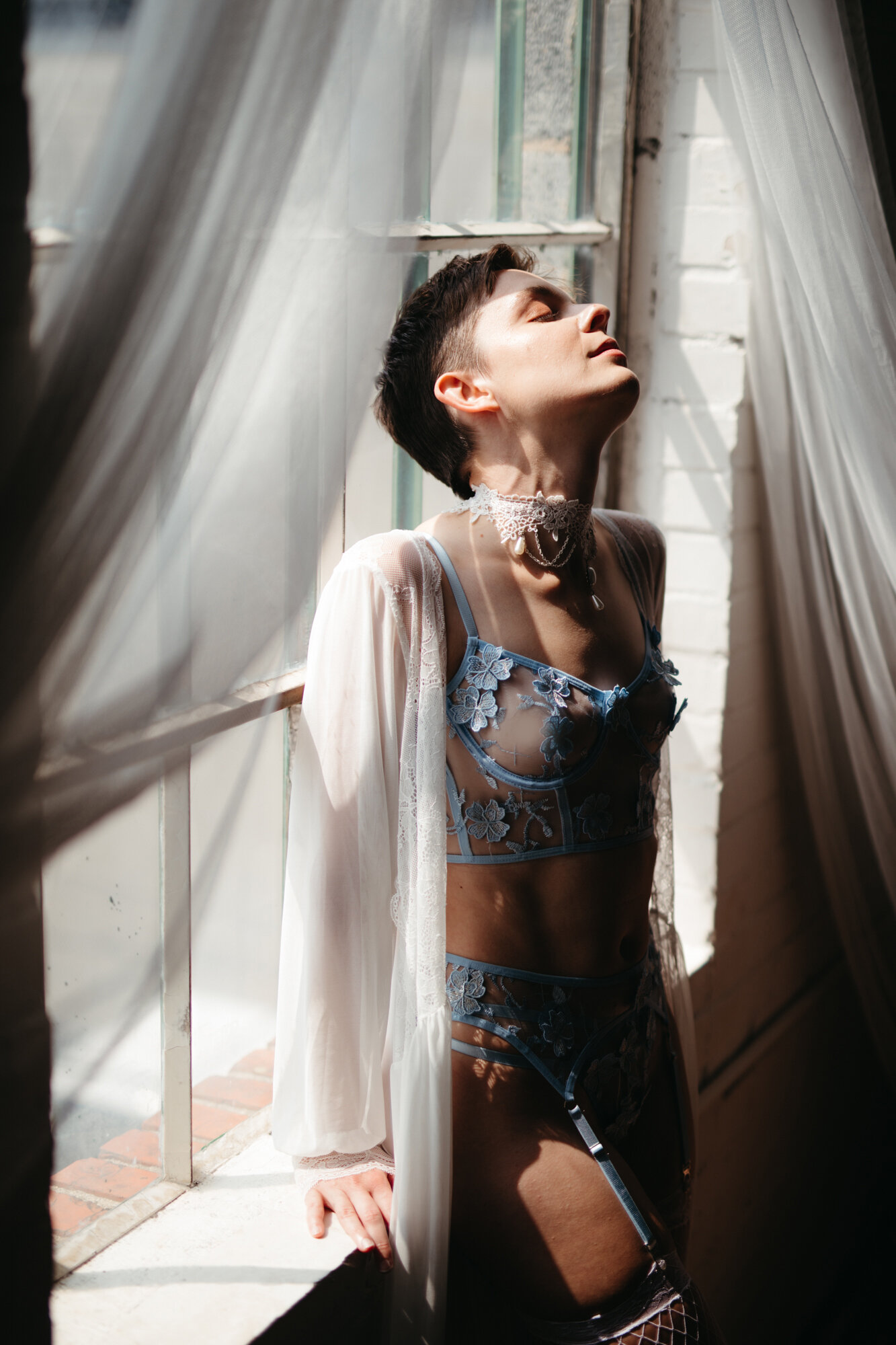 Genderqueer person with short hair sitting in a window wearing blue lingerie set