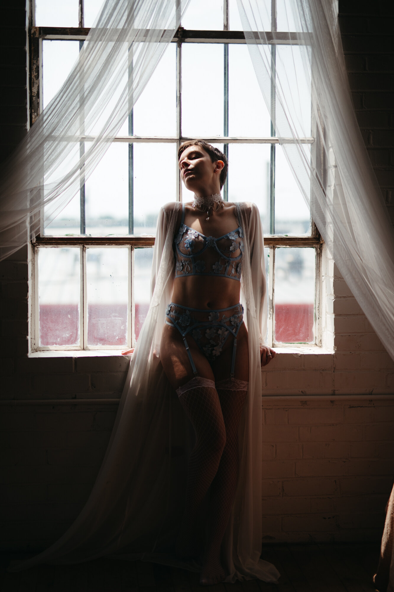 Genderqueer person wearing victorian style blue lingerie set silhouetted by a large window 