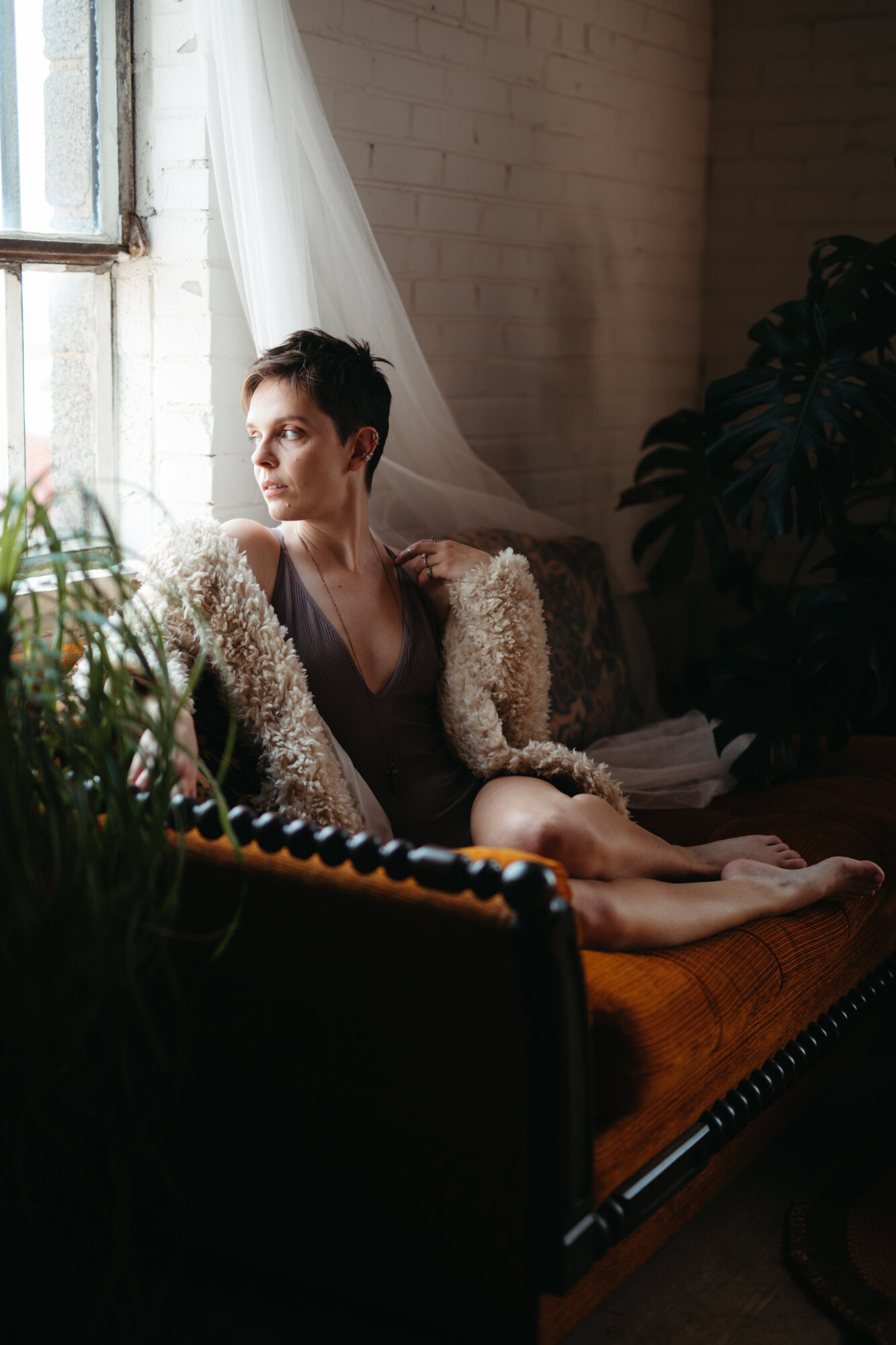 70's style boudoir photo of a person with short brown hair wearing a fuzzy coat sitting on a dark orange and brown couch.