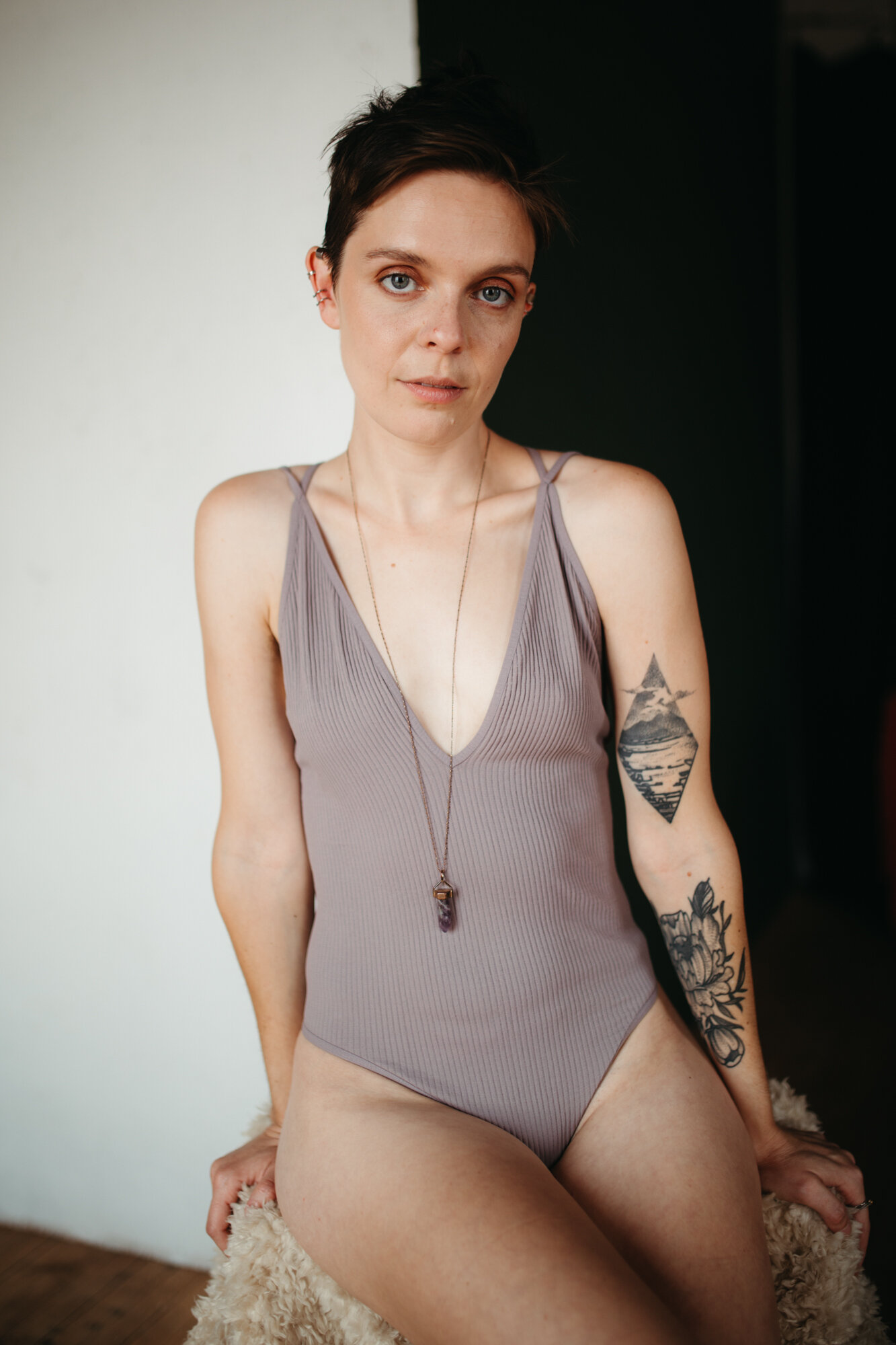 Queer boudoir photo of a person with tattoos in gray bodysuit with short brown hair looking directly into the camera