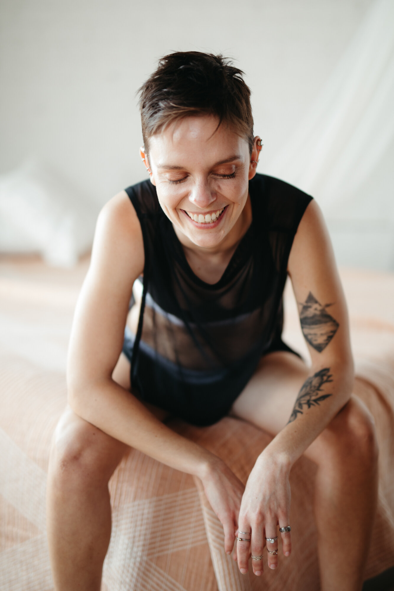 Queer masculine boudoir photo of person with short hair sitting on the edge of a bed laughing