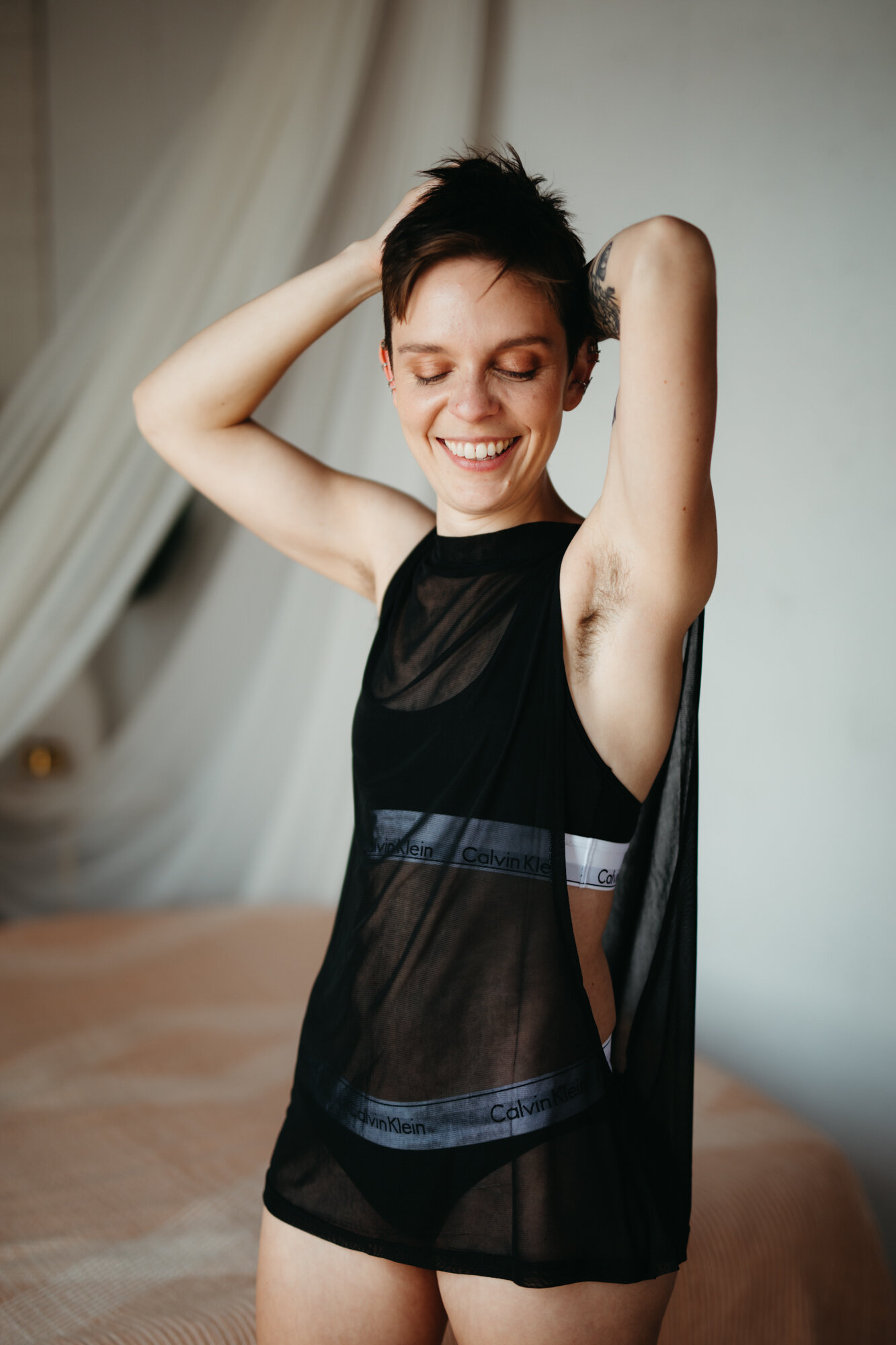 Queer joyous boudoir photo of person smiling with hands behind their head