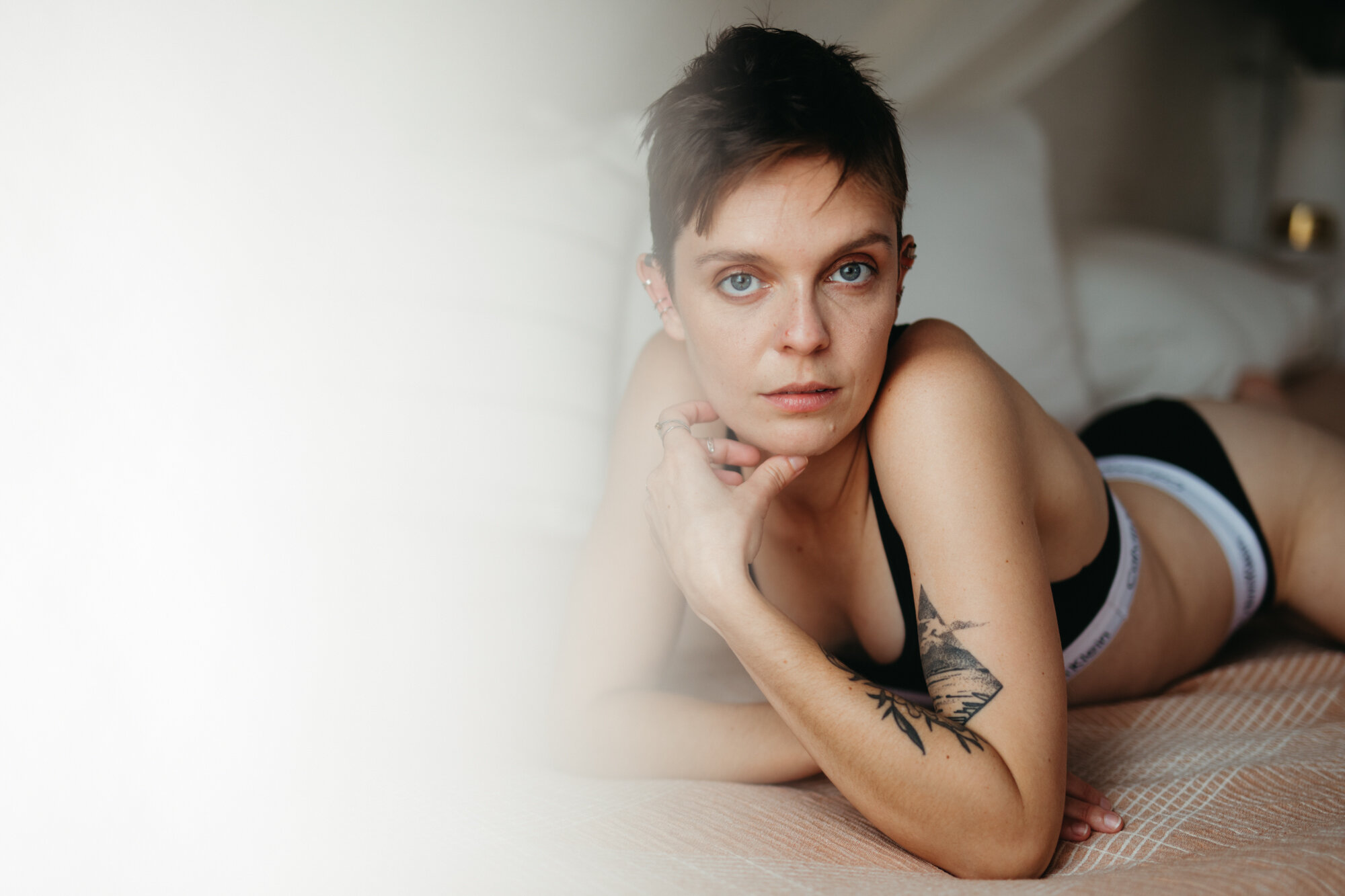 Androgynous boudoir photo of person with short brown hair laying on bed and looking into the camera